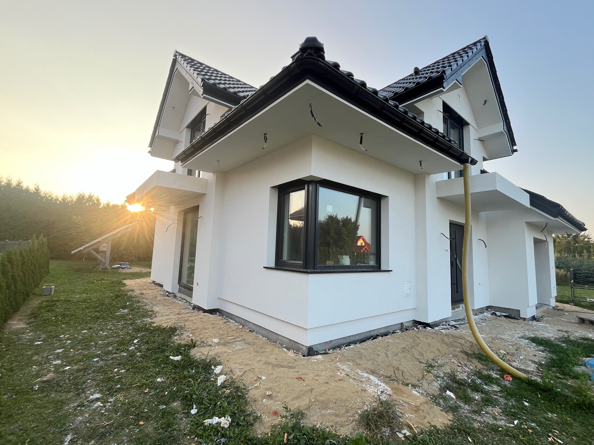 Nowoczesny dom jednorodzinny z białymi ścianami i czarnym dachem, widok z boku, teren wokół domu częściowo pokryty piaskiem, widoczne ślady prac budowlanych, żółty wąż leżący na ziemi, zachodzące...