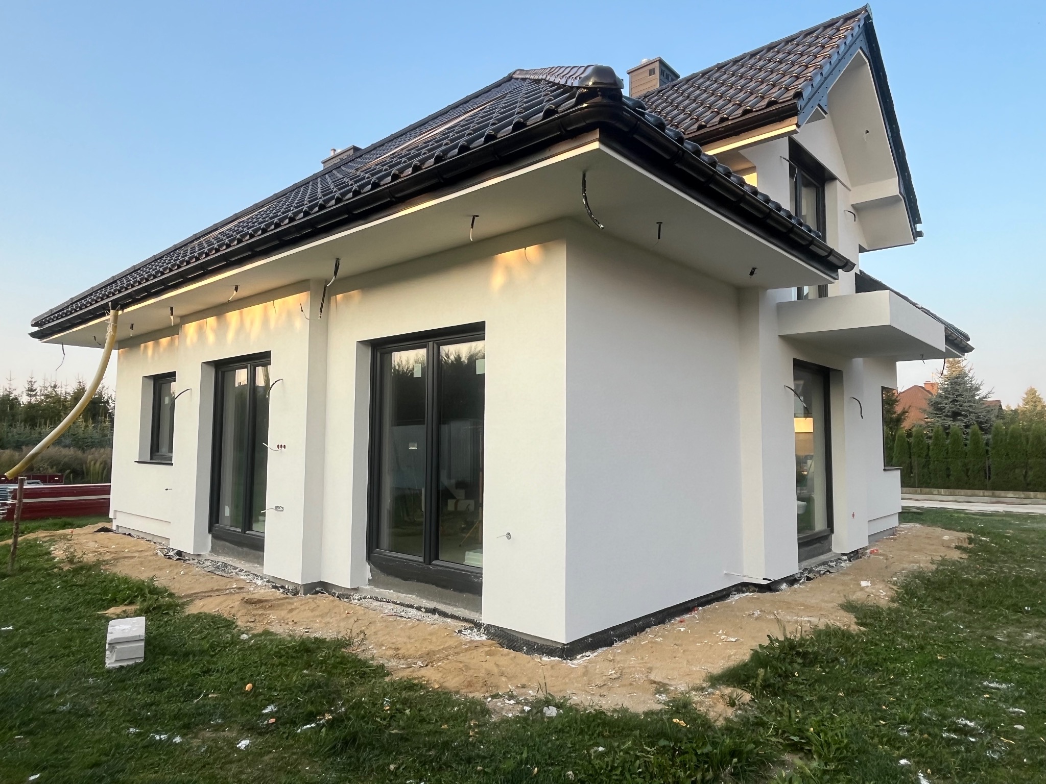Nowoczesny dom jednorodzinny z białymi ścianami elewacyjnymi i czarną dachówką, widoczne przygotowania do dalszych prac wykończeniowych wokół budynku.
