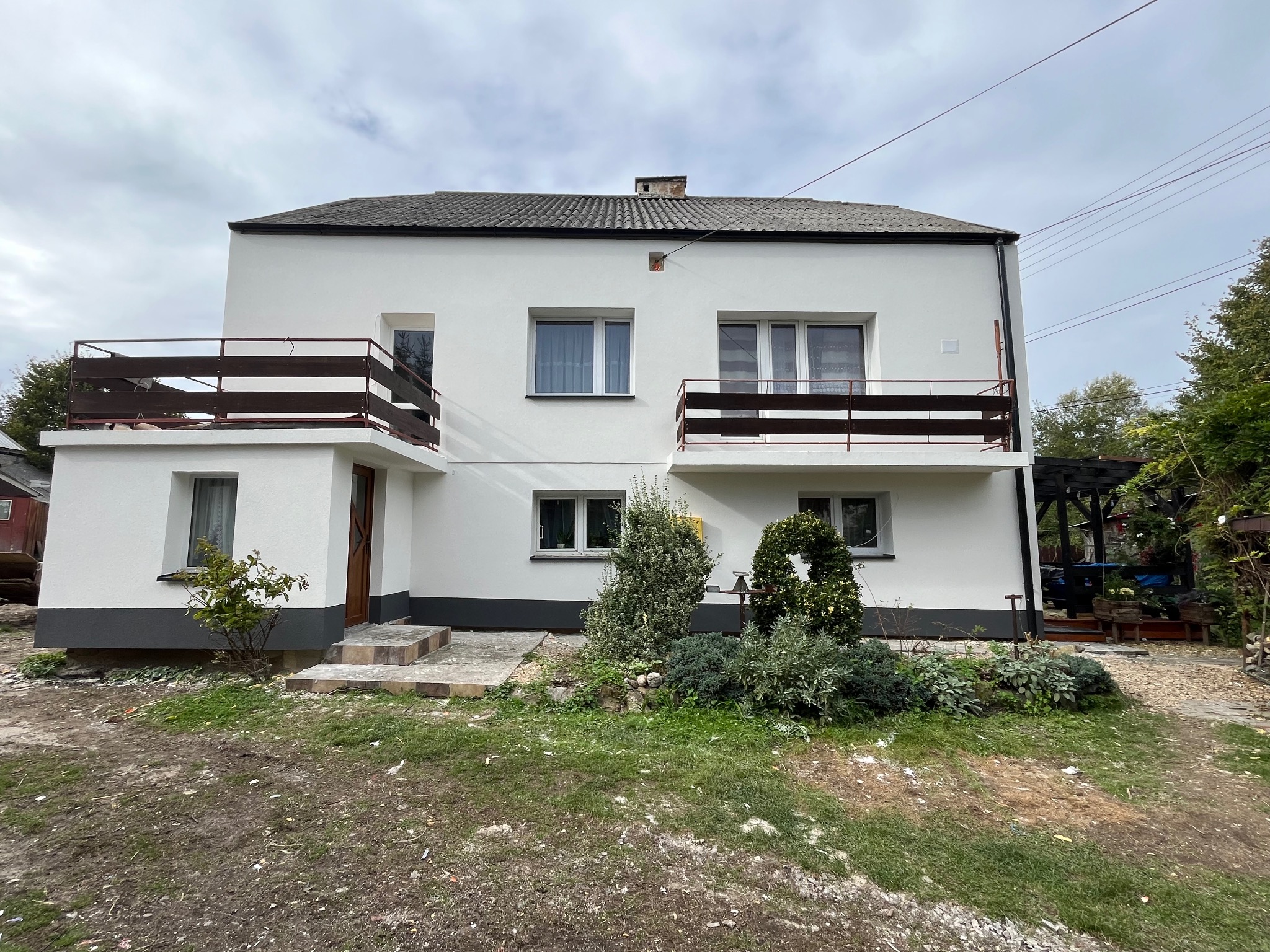 Dwukondygnacyjny dom z białymi ścianami i ciemnymi balkonami, widok z poziomu gruntu na front budynku, szare wykończenie cokołu, zieleń w ogrodzie.