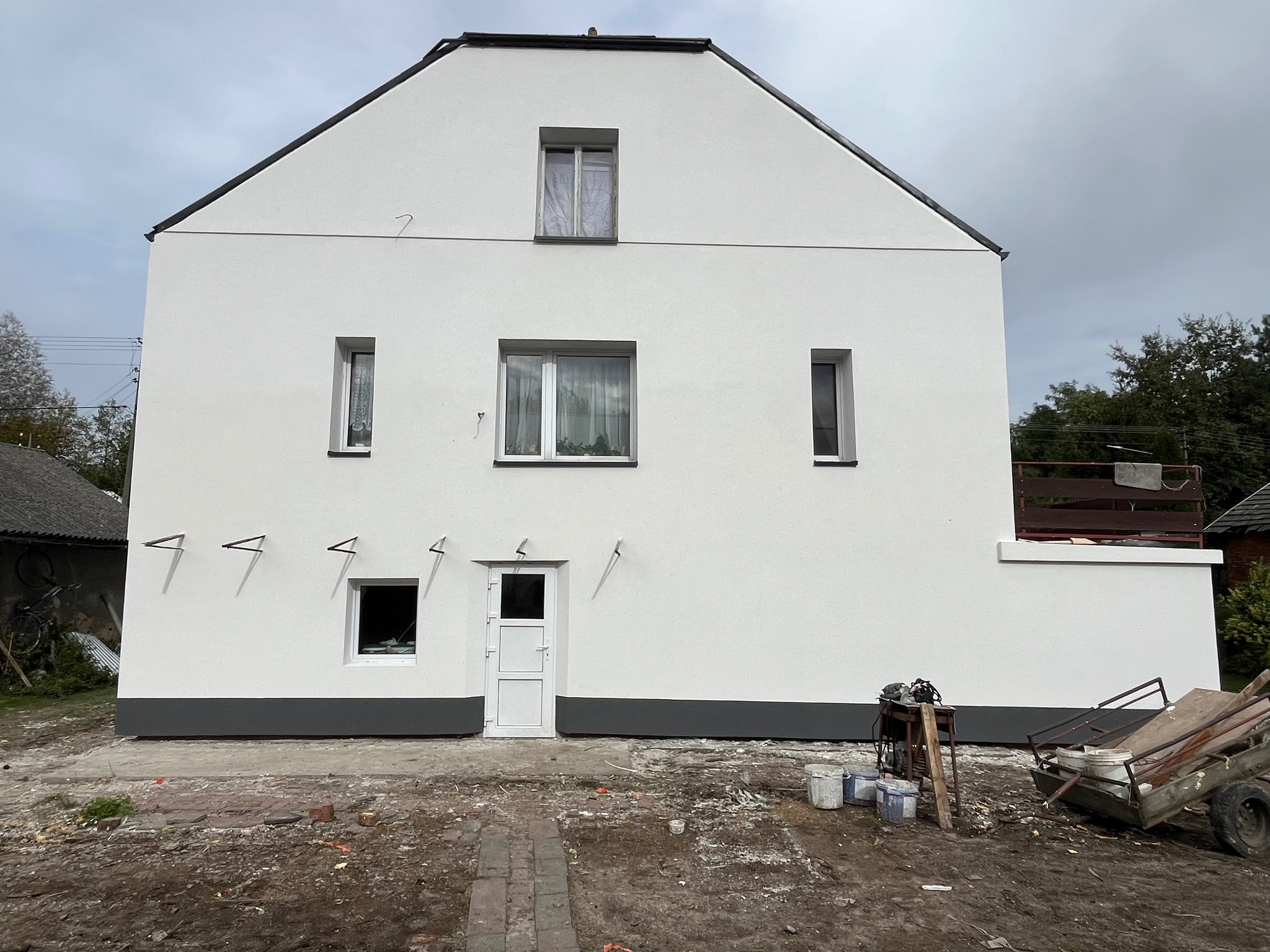 Świeżo otynkowany, biały dom jednorodzinny z szarym cokołem, widoczne ślady prac budowlanych na posesji, drabiny na elewacji, pochmurne niebo.