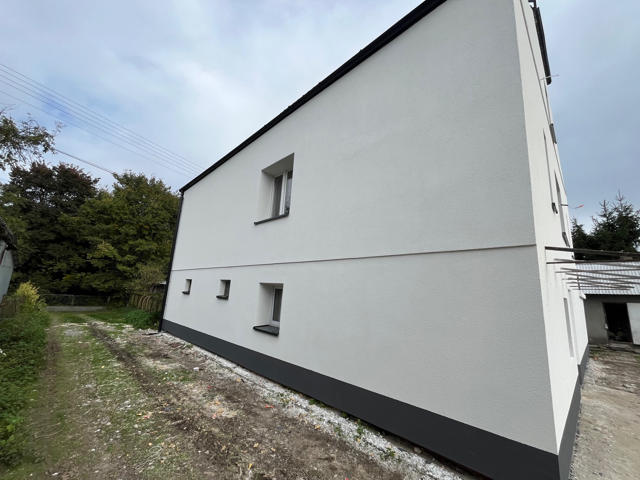 Nowo otynkowana i pomalowana na biało elewacja budynku z ciemnoszarą podmurówką i obramowaniami okien, widoczna z perspektywy ziemi w pochmurny dzień.