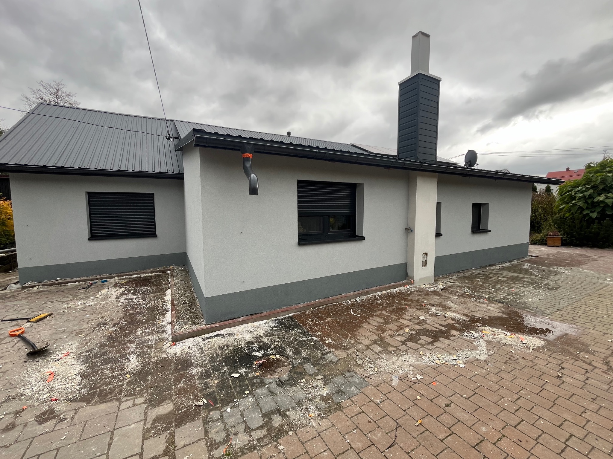 Dom jednorodzinny po tynkowaniu elewacji na szaro, z ciemnoszarą podmurówką i ciemnym dachem z blachy trapezowej, teren wokół domu częściowo wybrukowany kostką, widoczne resztki materiałów...