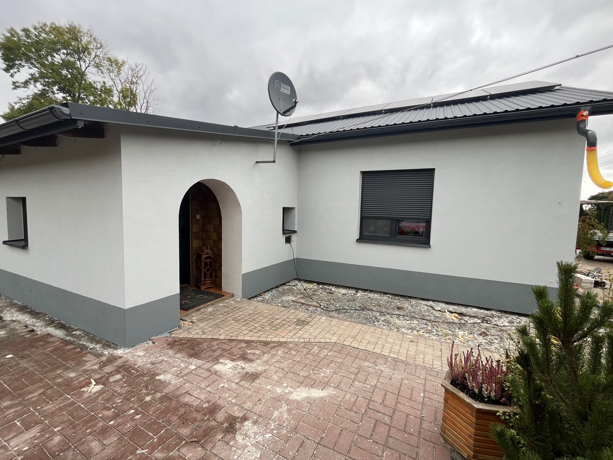 Wykończony dom jednorodzinny z jasną elewacją, szarą podmurówką i nową kostką brukową na podjeździe, widoczny także fragment dachu z panelami solarnymi i anteną satelitarną.