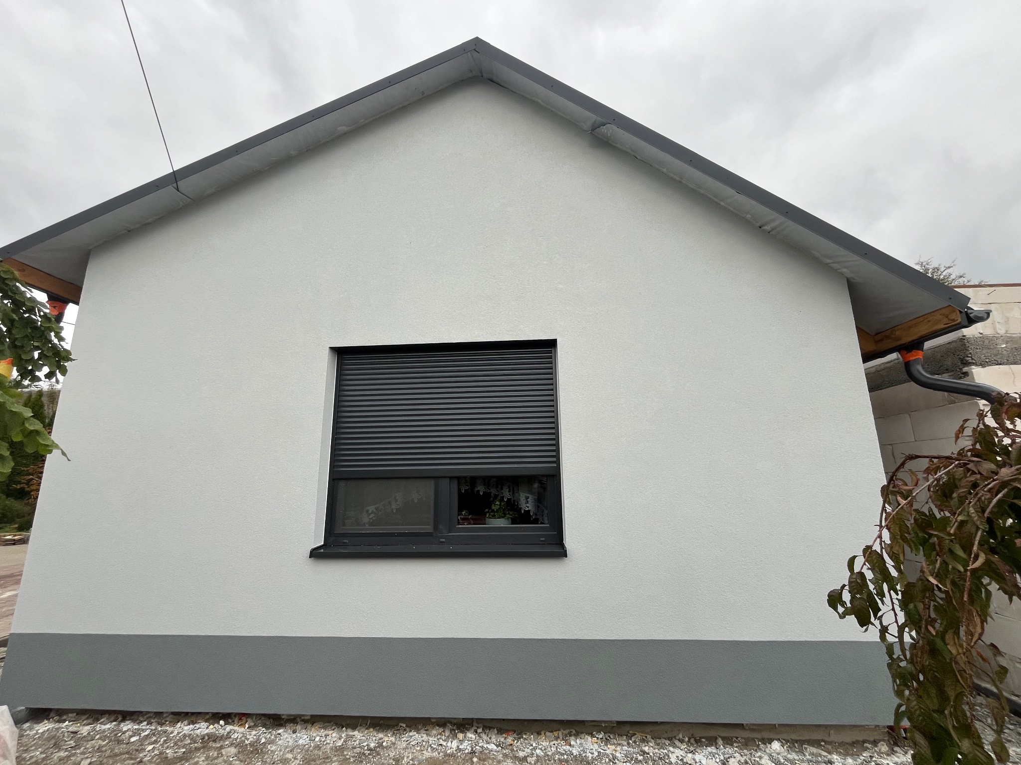 Świeżo otynkowana elewacja domu jednorodzinnego w odcieniach szarości i bieli, z widocznym oknem z roletą zewnętrzną, wykonana w pochmurny dzień.