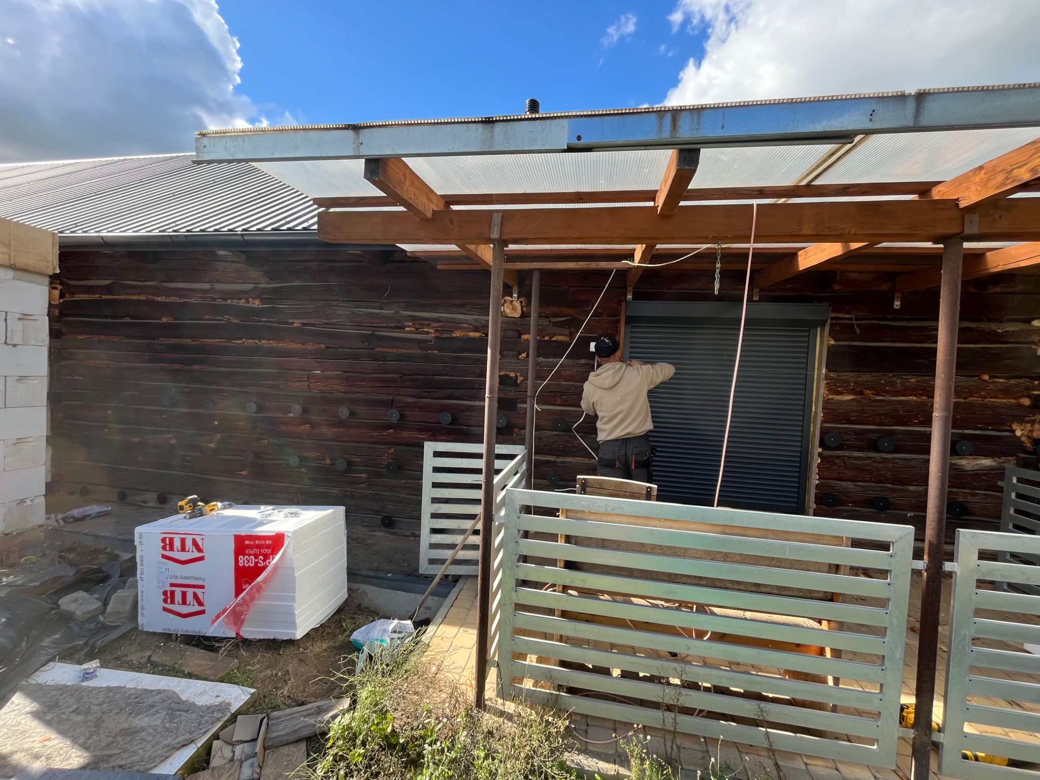 Prace wykończeniowe przy drewnianym domu z zadaszeniem: montaż rolet zewnętrznych, widoczne paczki styropianu NTB i częściowo zbudowany płot.