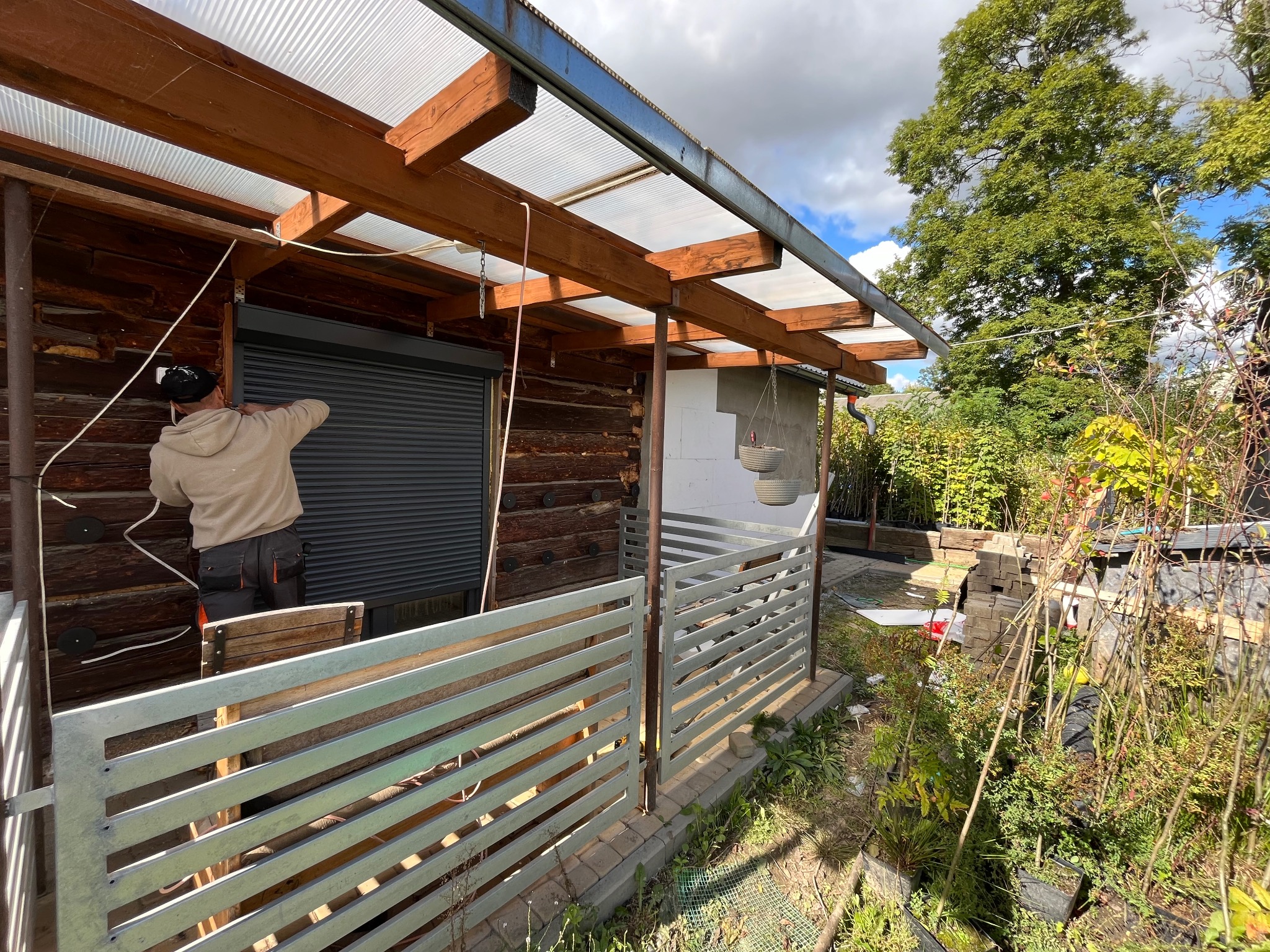 Pracownik w beżowej bluzie montuje roletę zewnętrzną na drewnianej ścianie budynku z widoczną konstrukcją zadaszenia z poliwęglanu i ogrodzeniem z metalowych sztachet.