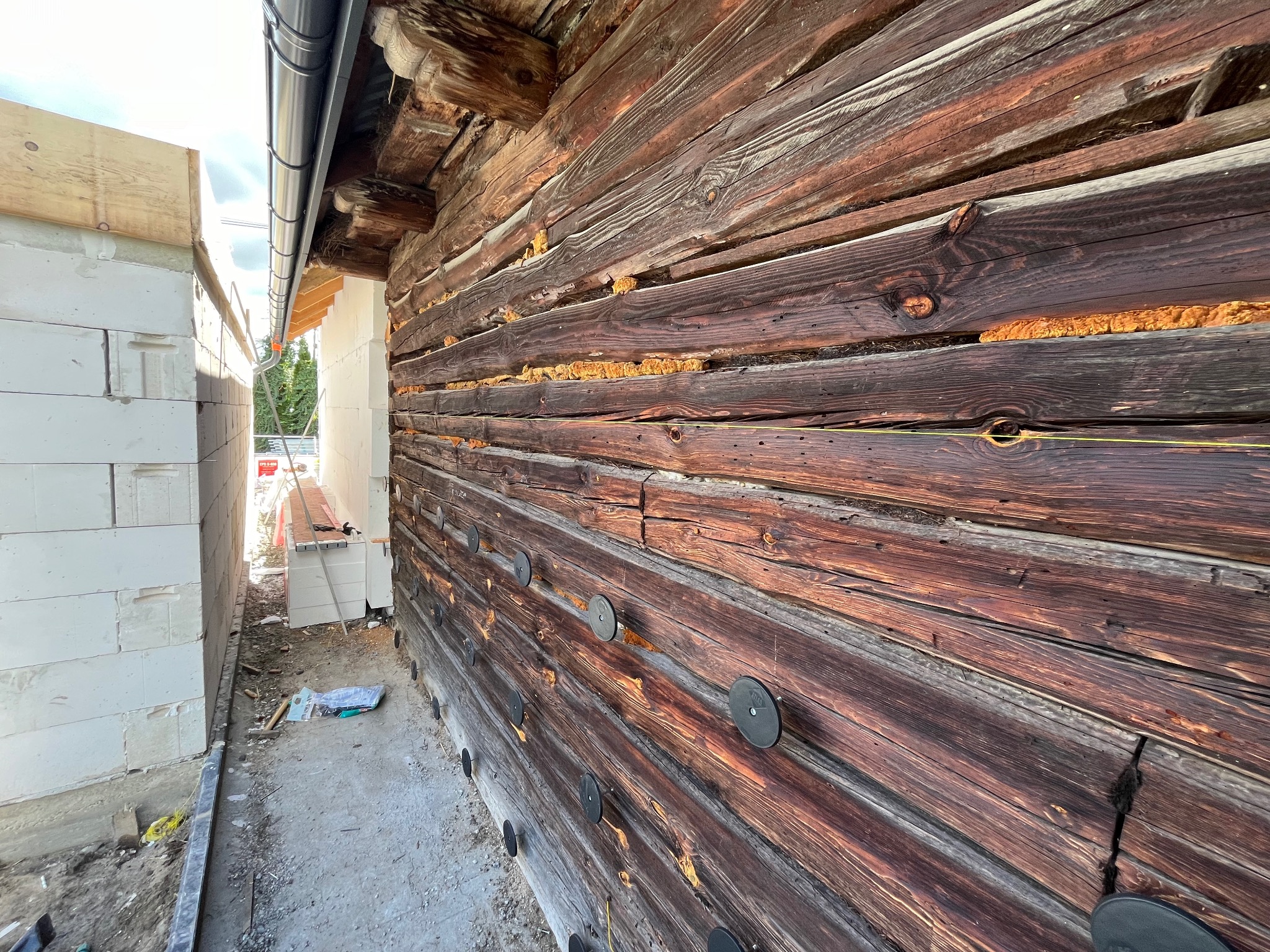 Fragment drewnianej ściany domu z widocznymi szczelinami wypełnionymi pianką izolacyjną i zamocowanymi okrągłymi elementami mocującymi, sąsiadujący z fragmentem ściany z białych bloczków.