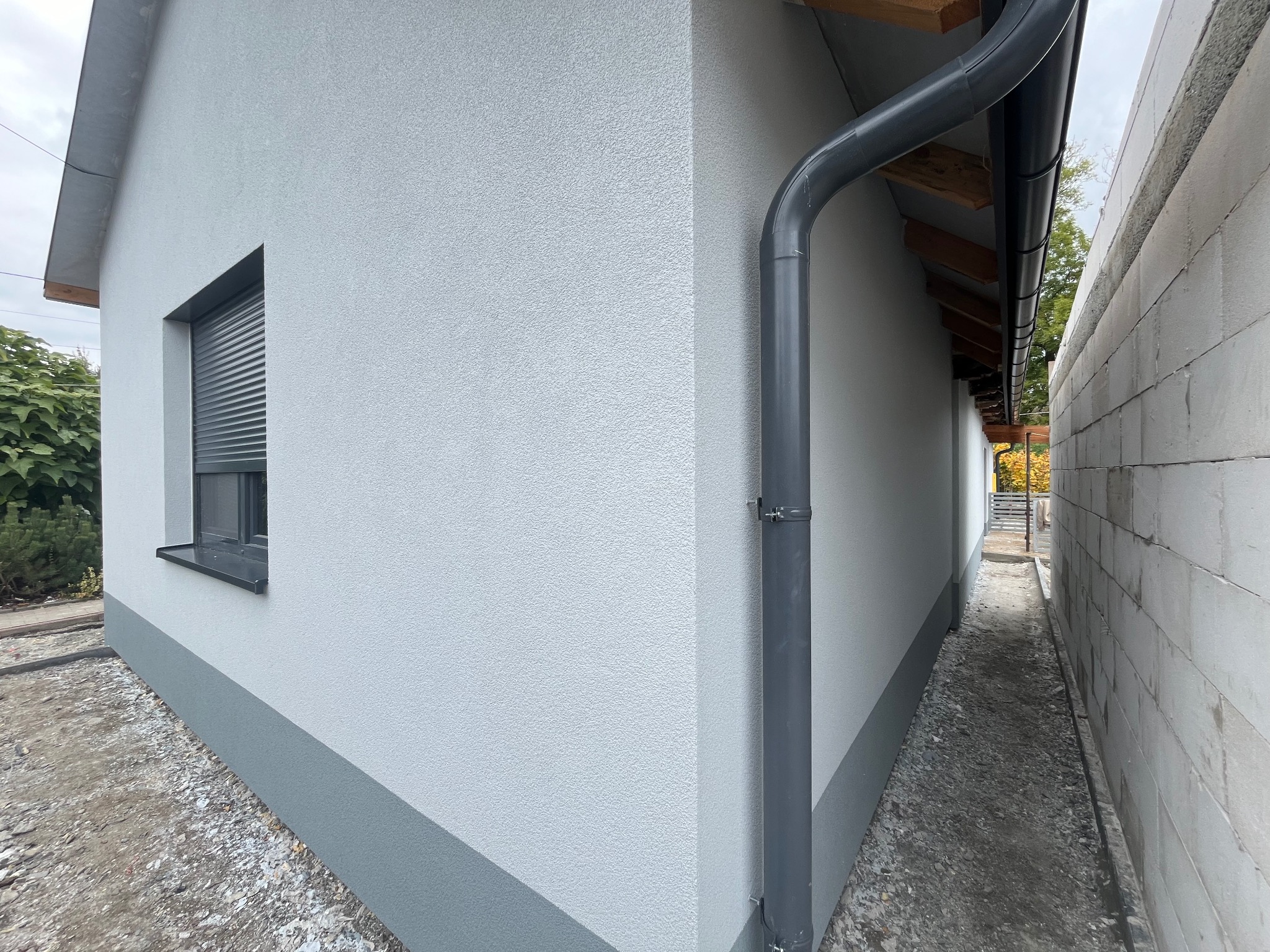 Jasnoszara elewacja domu jednorodzinnego z widoczną rynną, oknem z roletą zewnętrzną i fragmentem ścieżki wzdłuż budynku, wykończona dwoma odcieniami szarości.