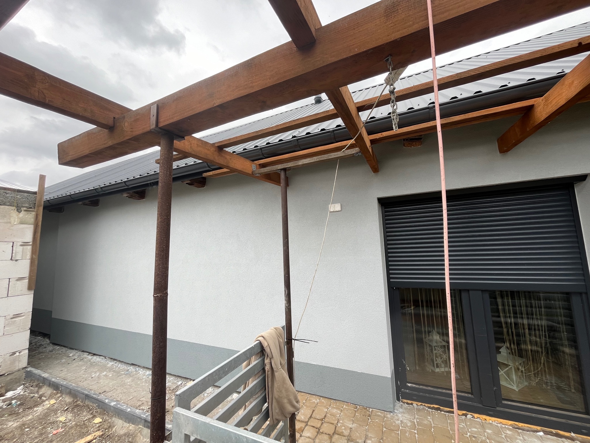 Świeżo otynkowana elewacja domu jednorodzinnego z drewnianą konstrukcją zadaszenia, widoczne rusztowanie i okno z roletami zewnętrznymi, w tle pochmurne niebo.