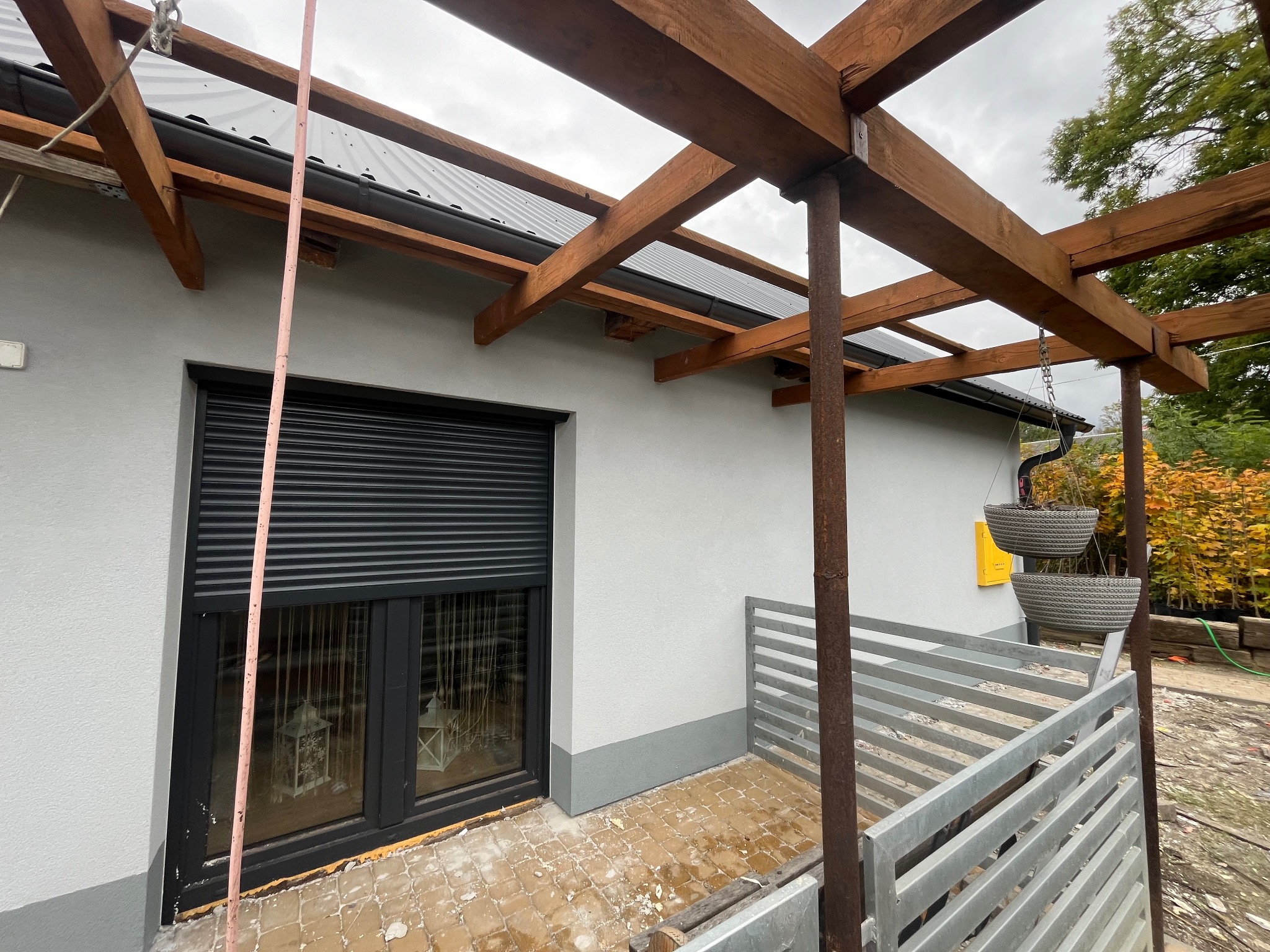 Szara elewacja domu z drewnianą konstrukcją zadaszenia tarasu w trakcie budowy, widoczny fragment okna z roletą zewnętrzną i fragment terenu z kostką brukową.