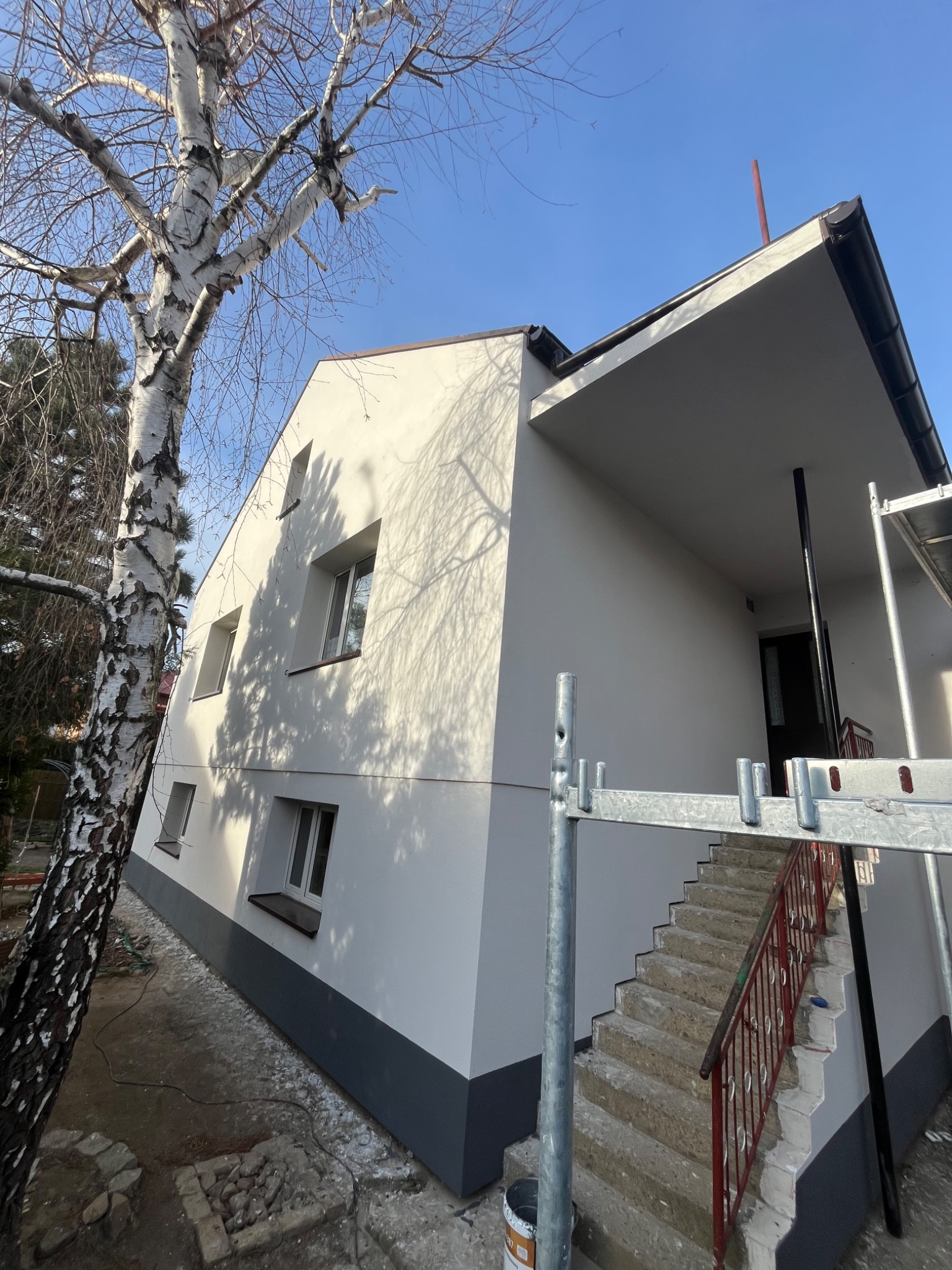 Świeżo pomalowana elewacja domu z widocznym rusztowaniem, dolna część ściany w kolorze szarym, a górna biała, obok stoi brzoza bez liści i widać cień rzucany przez gałęzie na ścianę.