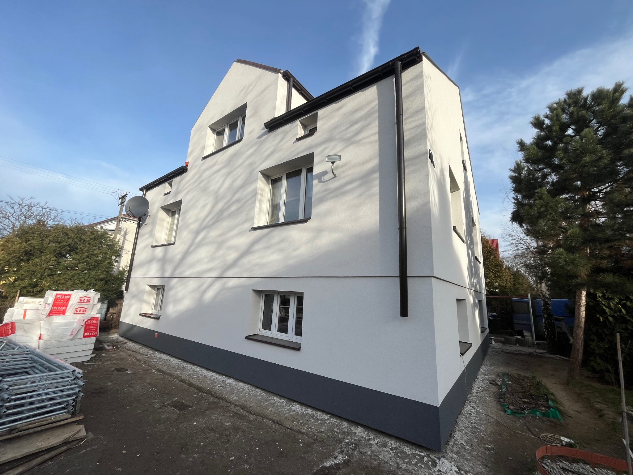 Nowo otynkowany, biały dom z szarą podmurówką, widok z zewnątrz, z boku leżą sterty styropianu i rusztowanie, błękitne niebo w tle.