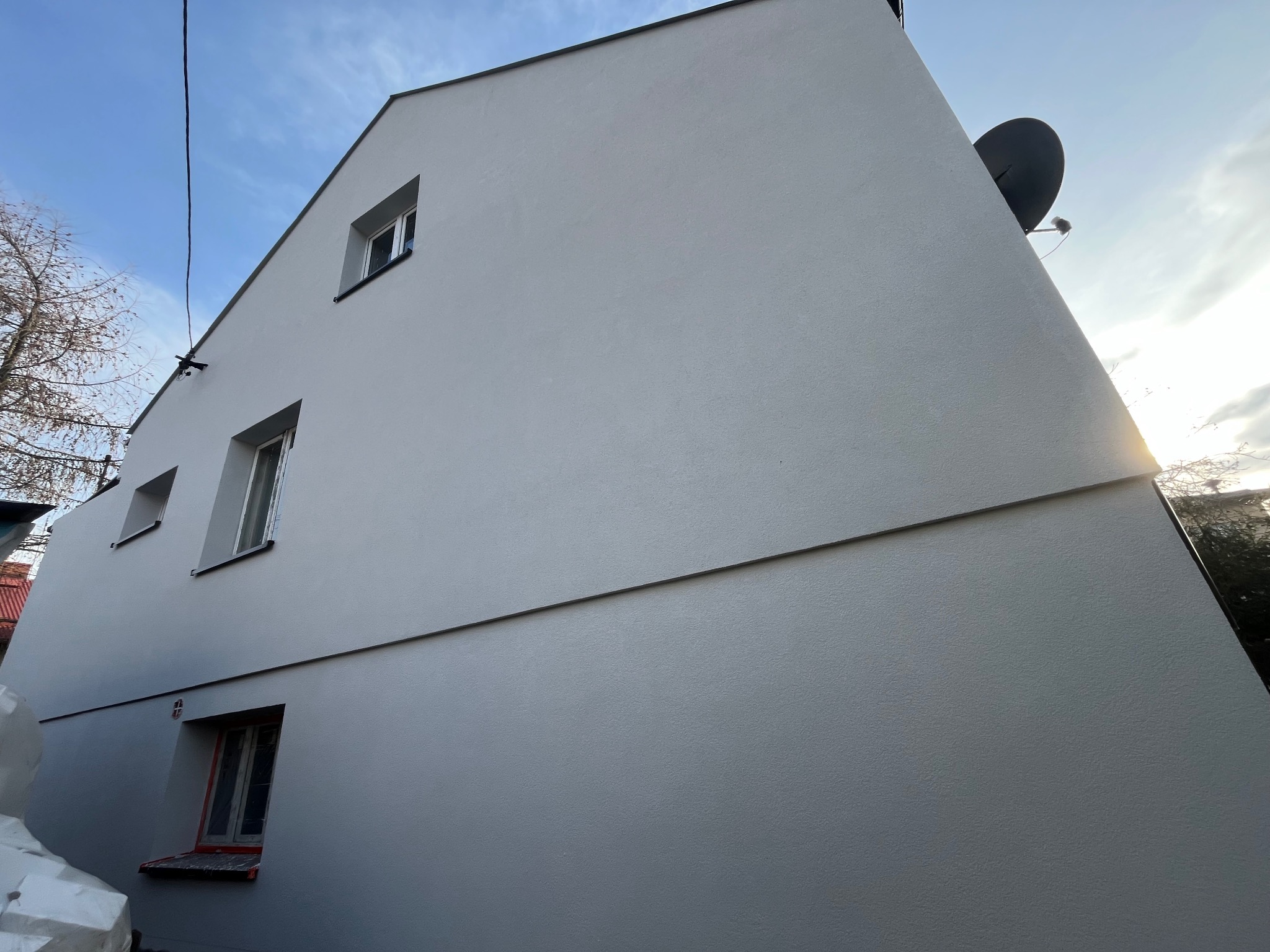 Świeżo otynkowana ściana domu jednorodzinnego z zamontowaną anteną satelitarną, widok z dołu na szarą elewację w słoneczny dzień.
