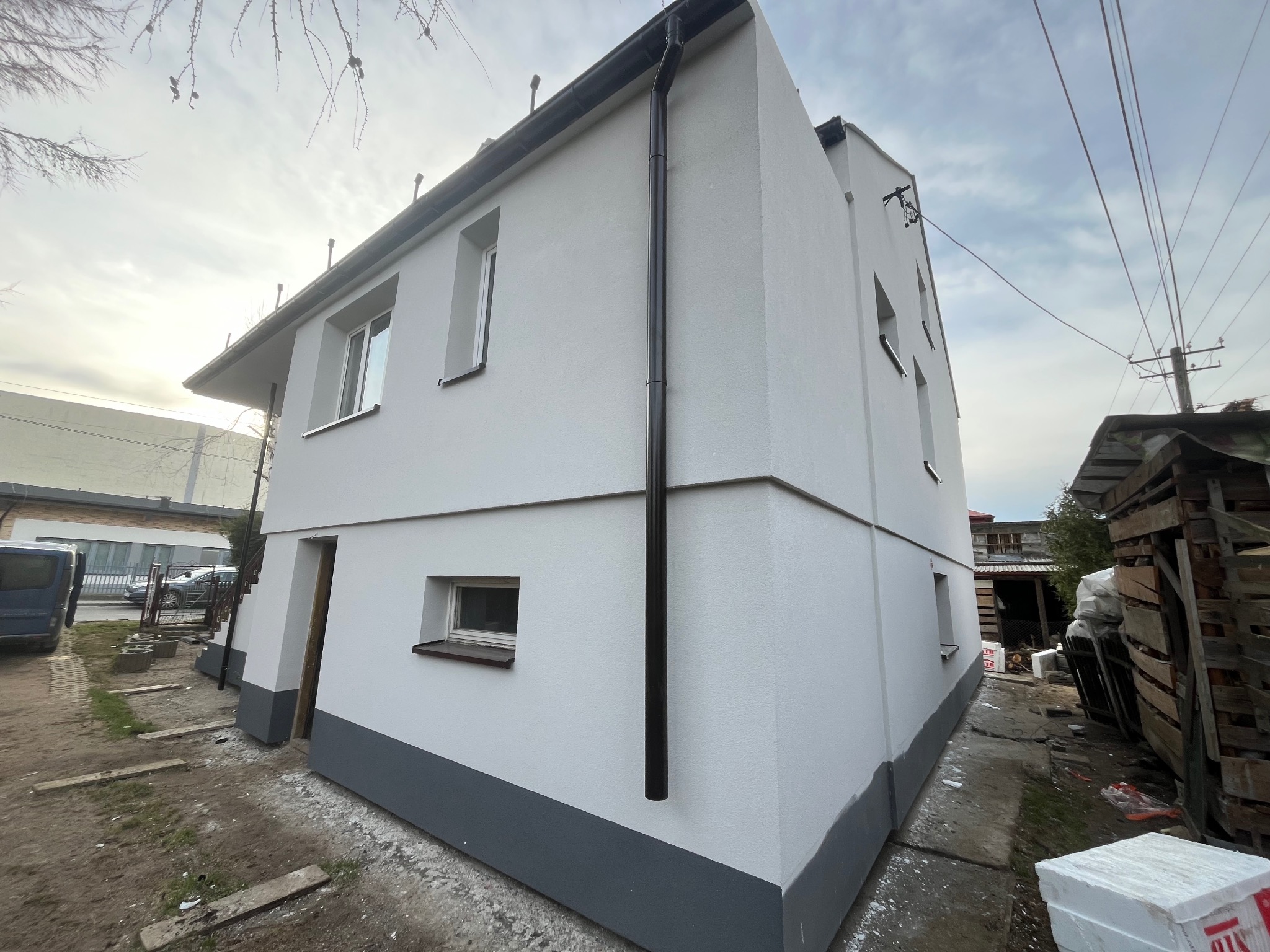 Dwupiętrowy budynek z nową, jasnoszarą elewacją i ciemnoszarą podmurówką, widoczny z boku, z rynną spustową, oknami z brązowymi ramami, częściowo ocieplony styropianem na zewnątrz, na tle...