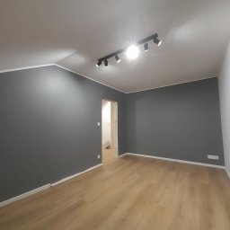 Jakub Zmuda - Wnętrze pokoju po remoncie: świeżo pomalowane szare ściany z białymi listwami przypodłogowymi i nową podłogą z paneli, widoczna czarna lampa sufitowa z kilkoma reflektorami.