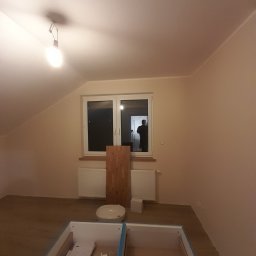 Jakub Zmuda - Pokój na poddaszu w trakcie prac wykończeniowych: widoczna surowa podłoga, świeżo pomalowane ściany, okno z widocznym odbiciem sylwetki, niedokończona szafka z szufladami na pierwszym planie...