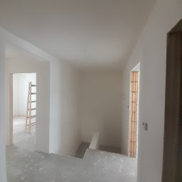 Jakub Zmuda - Surowe wnętrze w trakcie prac wykończeniowych: gładkie, białe ściany, szary betonowy podkład, widoczne cegły w ościeżnicy okiennej, drewniana drabina w tle.