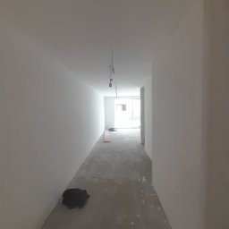 Jakub Zmuda - Korytarz w trakcie remontu: gładzie na ścianach, surowa podłoga, wiszące przewody elektryczne, resztki materiałów budowlanych, widok w kierunku okna.