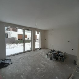 Jakub Zmuda - Wnętrze w trakcie prac wykończeniowych: widoczne puszki z farbą, zakurzona podłoga i ściany po wstępnym malowaniu, z widocznymi puszkami elektrycznymi. Na zewnątrz okien widoczny ośnieżony ogród.