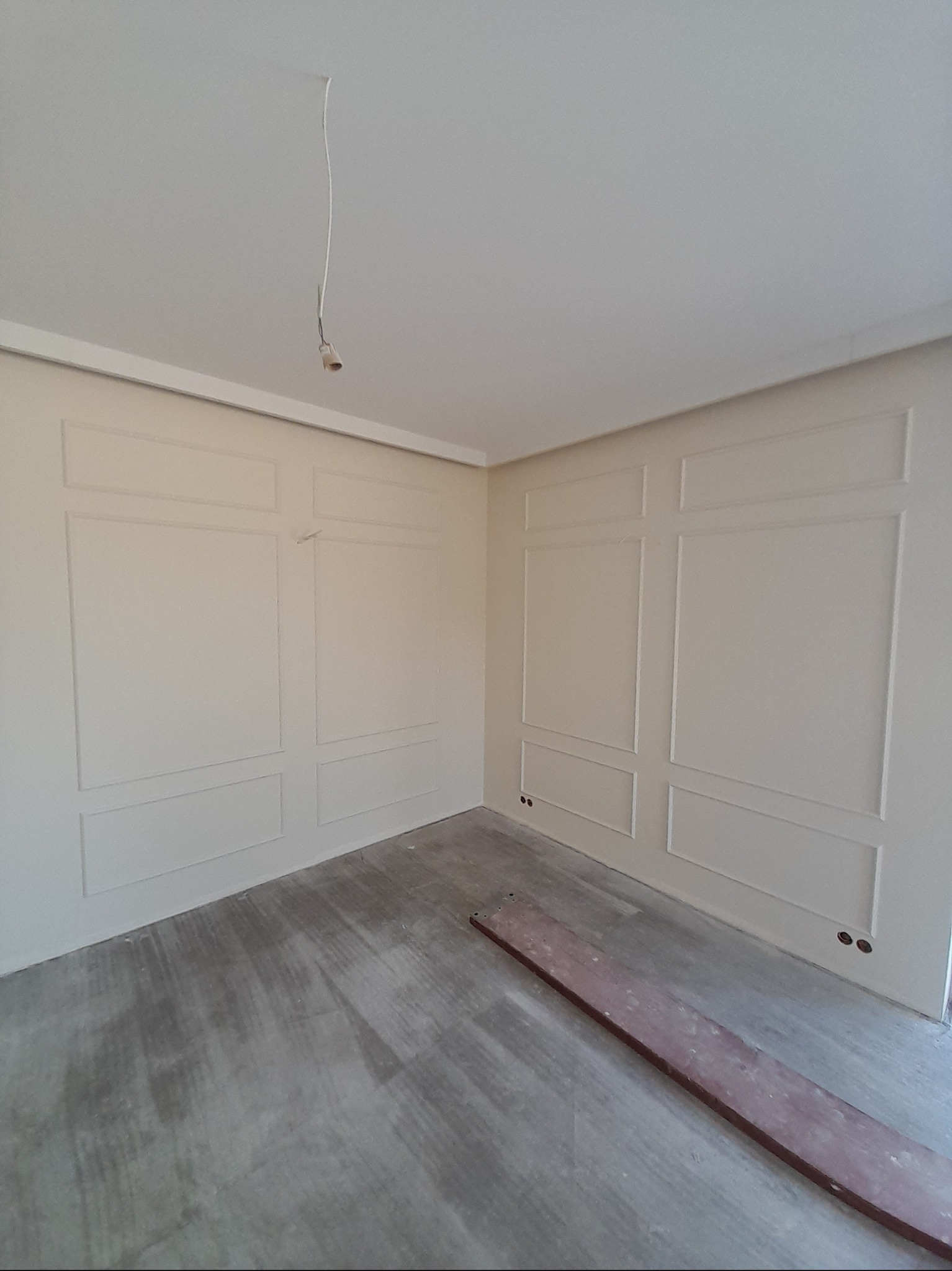 Pokój z dekoracyjnymi panelami ściennymi w odcieniach beżu. Na podłodze widoczna deska, a z sufitu zwisa kabel. Ściany wykończone w stylu klasycznym.