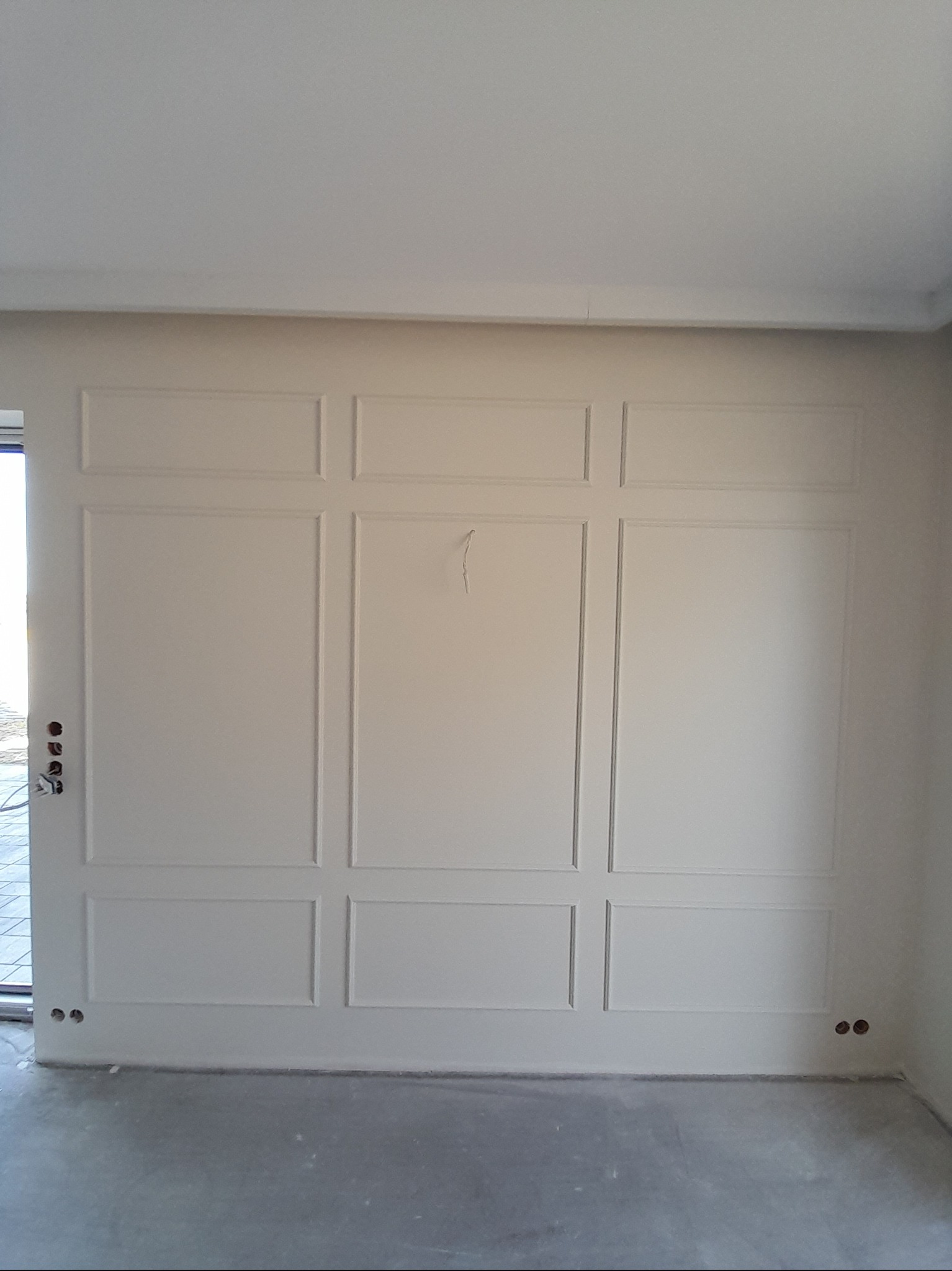 Wykończona ściana z dekoracyjnymi panelami w jasnym odcieniu, widoczne gniazdka elektryczne i fragment drzwi. Minimalistyczny design wnętrza.