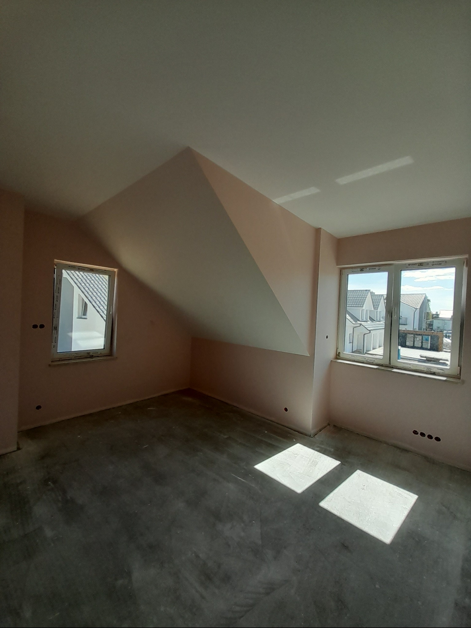 Poddasze po szpachlowaniu, przygotowane do malowania. Widoczne dwa okna z widokiem na zabudowę jednorodzinną, różowe ściany i szary betonowy podkład.