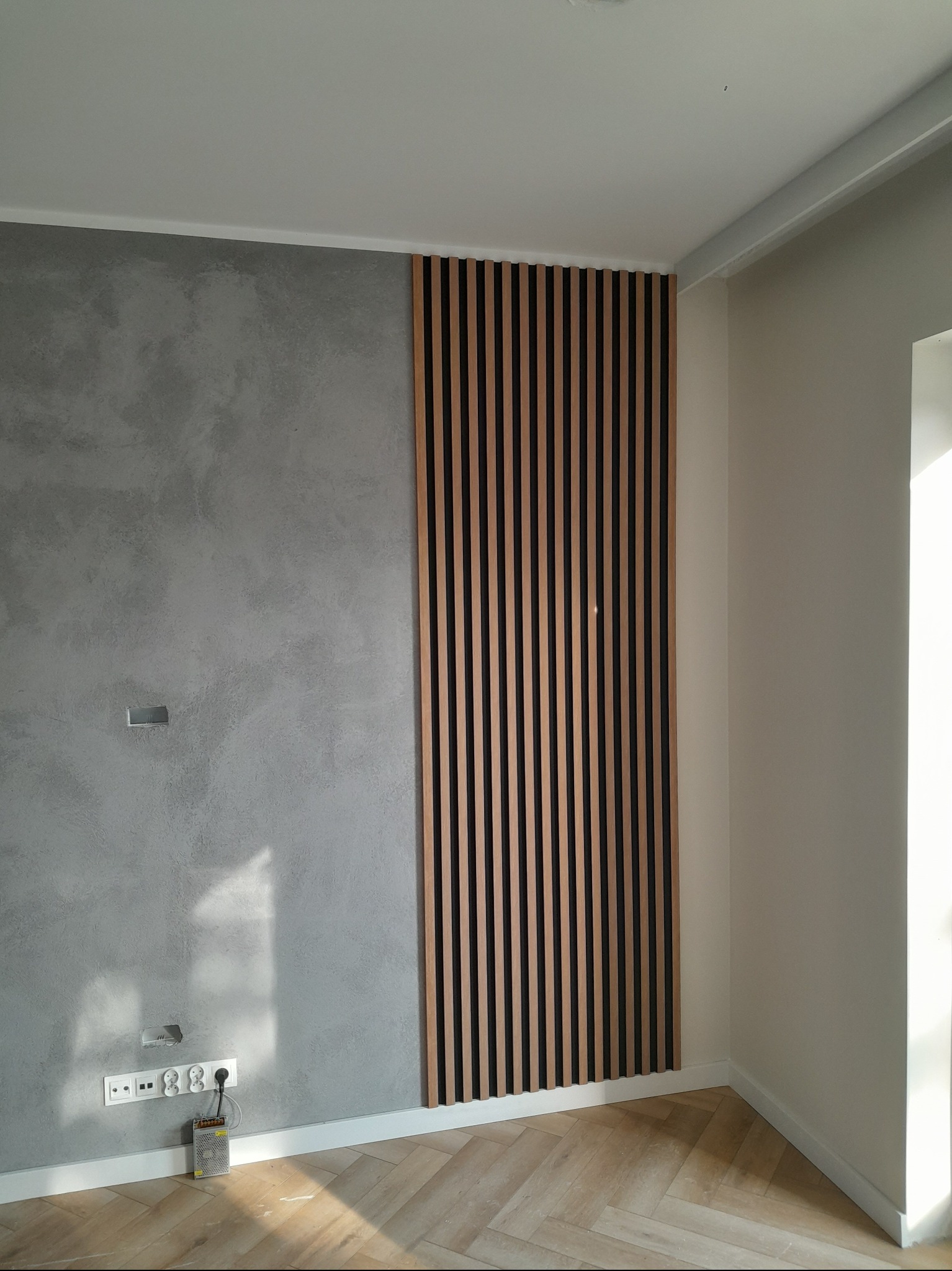 Nowoczesna ściana z dekoracyjną gładzią i drewnianymi lamelami w pionowym układzie, uzupełniona o listwę przypodłogową i parkiet w jodełkę.