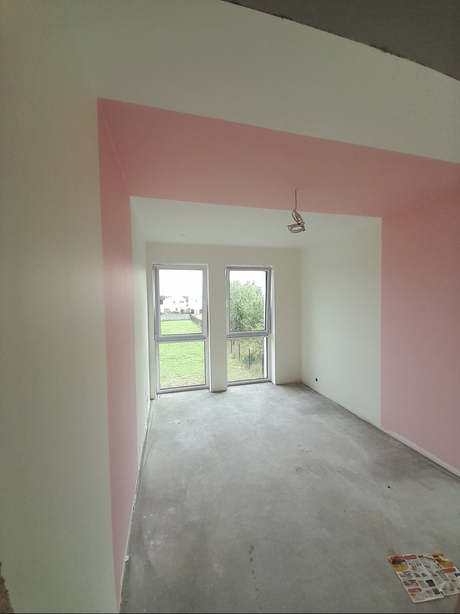 Wnętrze po malowaniu: białe ściany z różowymi akcentami wokół okien i sufitu. Surowa podłoga, dwa okna z widokiem na zieleń. Częściowo pomalowane ściany.