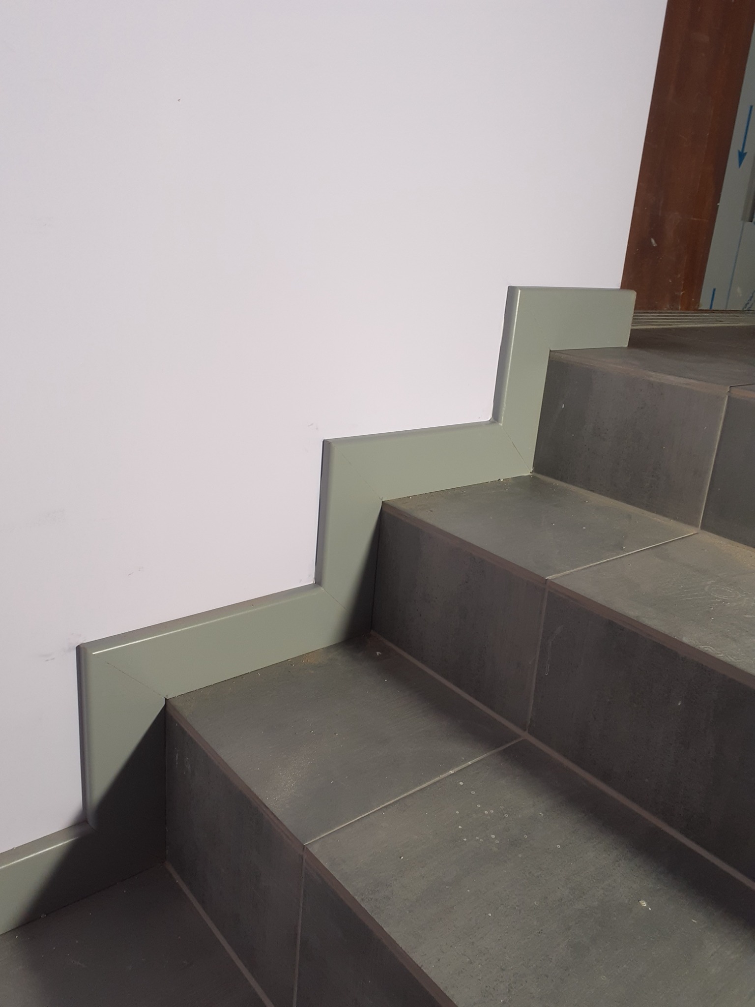 Szare płytki na schodach z dopasowaną kolorystycznie listwą przypodłogową przy białej ścianie, widok z dołu.