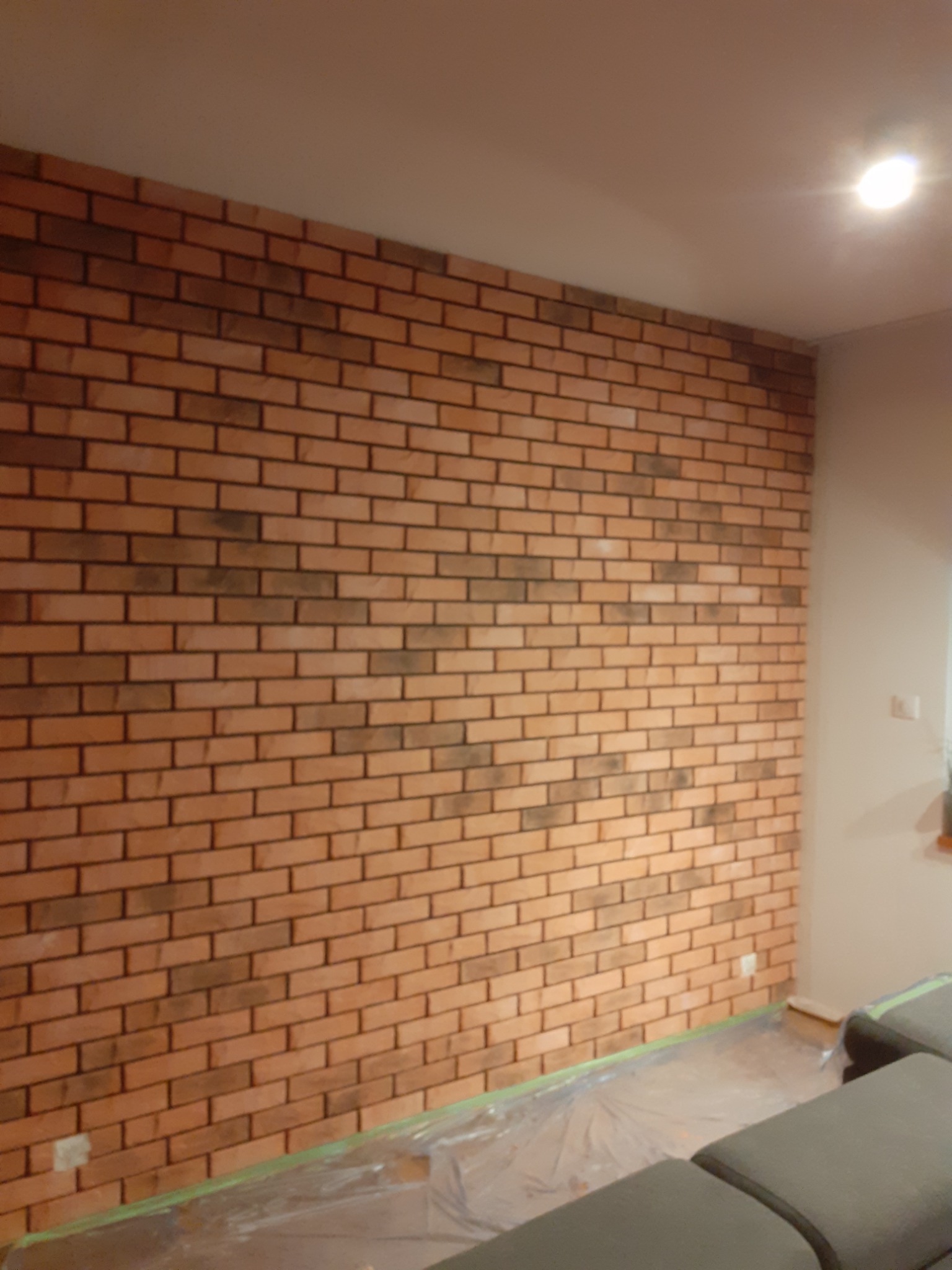 Ściana wykończona cegłą dekoracyjną w odcieniach czerwieni i brązu, widoczne zabezpieczone folią podłoże, lampa sufitowa w tle.