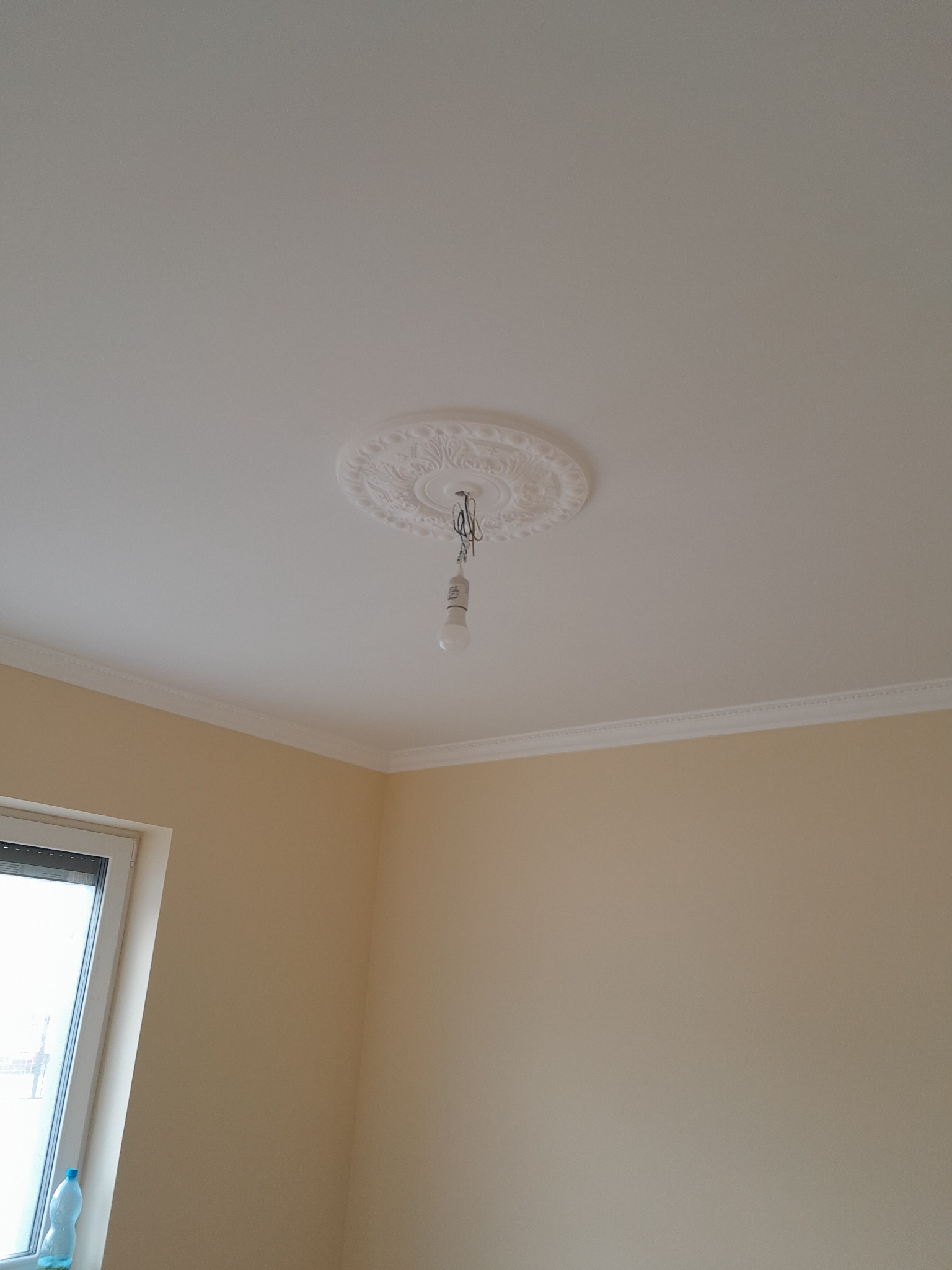 Pokój po remoncie: gładka, biała powierzchnia sufitu z dekoracyjną rozetą i wiszącą żarówką, kremowe ściany z listwami przypodłogowymi, okno z widoczną butelką wody.