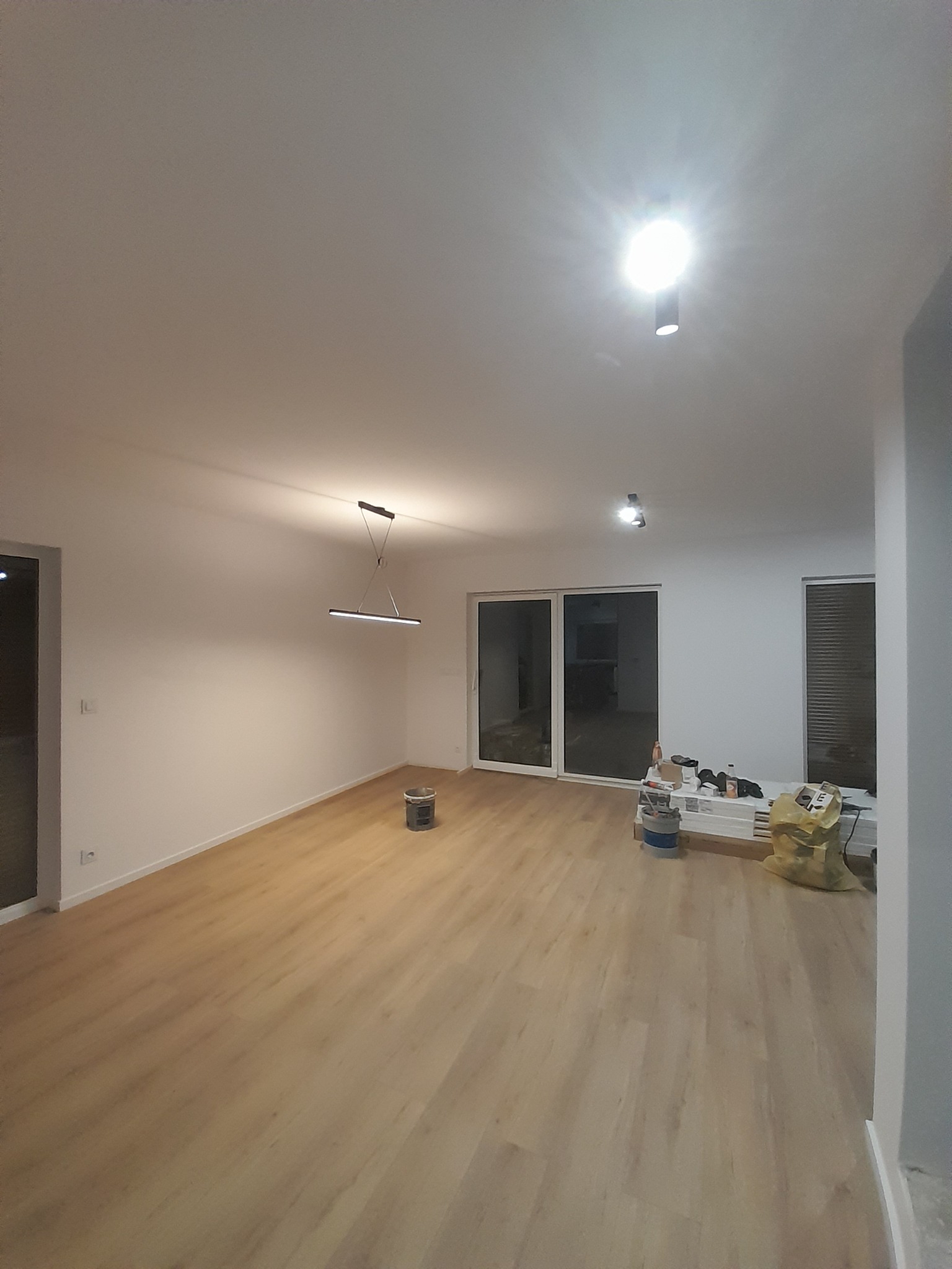 Jasne wnętrze po remoncie z panelami podłogowymi i świeżo pomalowanymi ścianami, widoczne przesuwne drzwi balkonowe, nowoczesna lampa wisząca i przygotowane materiały malarskie.