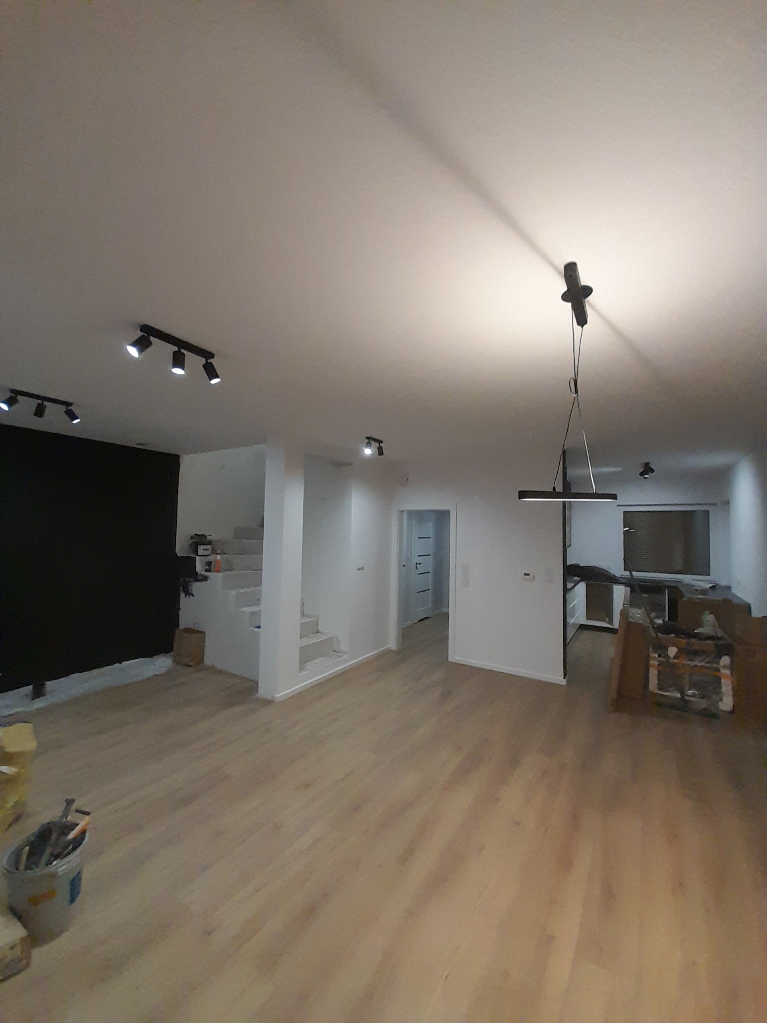 Przestronne wnętrze domu w trakcie remontu, z nową podłogą z paneli, czarną ścianą, białymi schodami i nowoczesnym oświetleniem sufitowym.