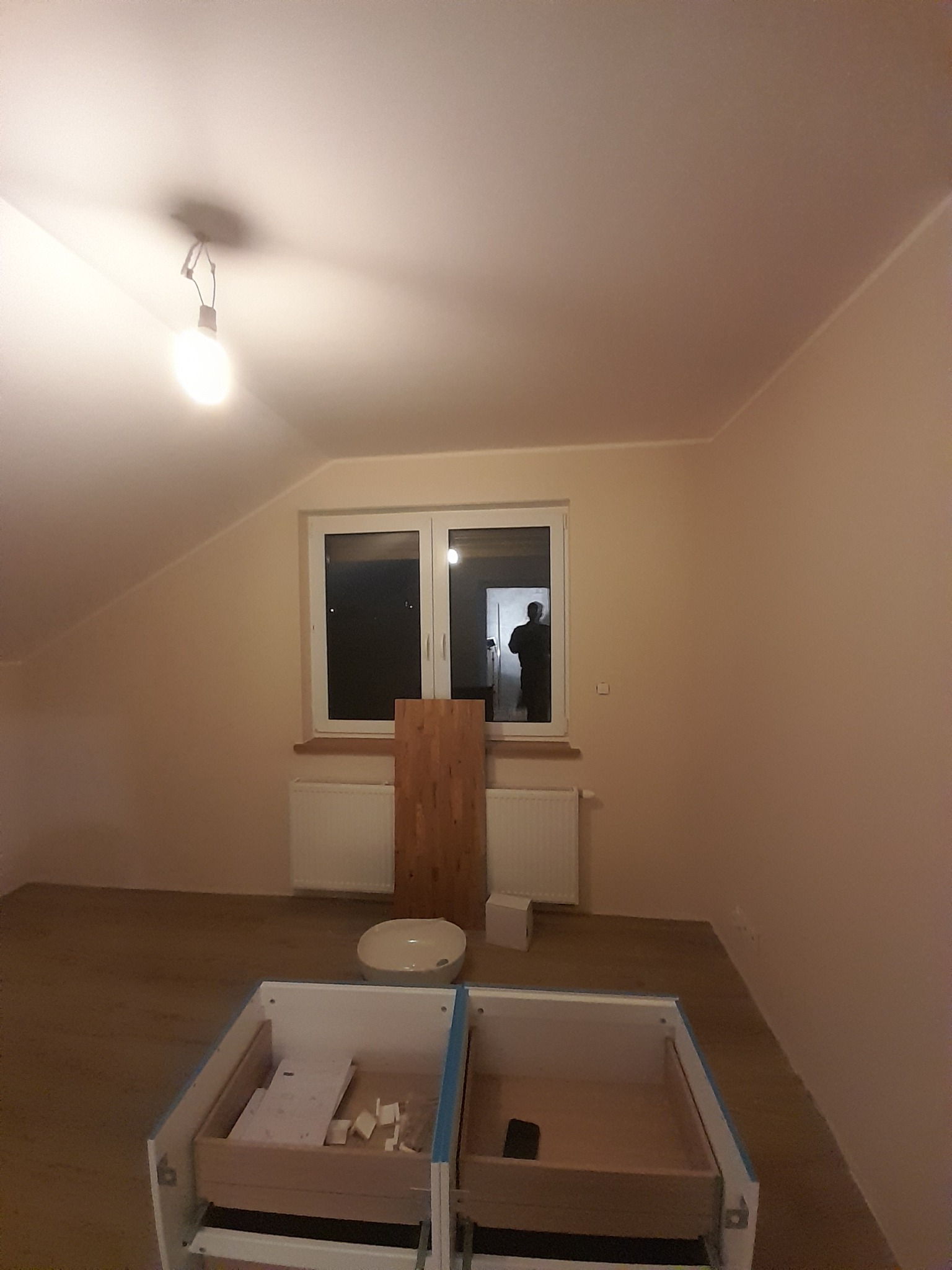 Pokój na poddaszu w trakcie prac wykończeniowych: widoczna surowa podłoga, świeżo pomalowane ściany, okno z widocznym odbiciem sylwetki, niedokończona szafka z szufladami na pierwszym planie...