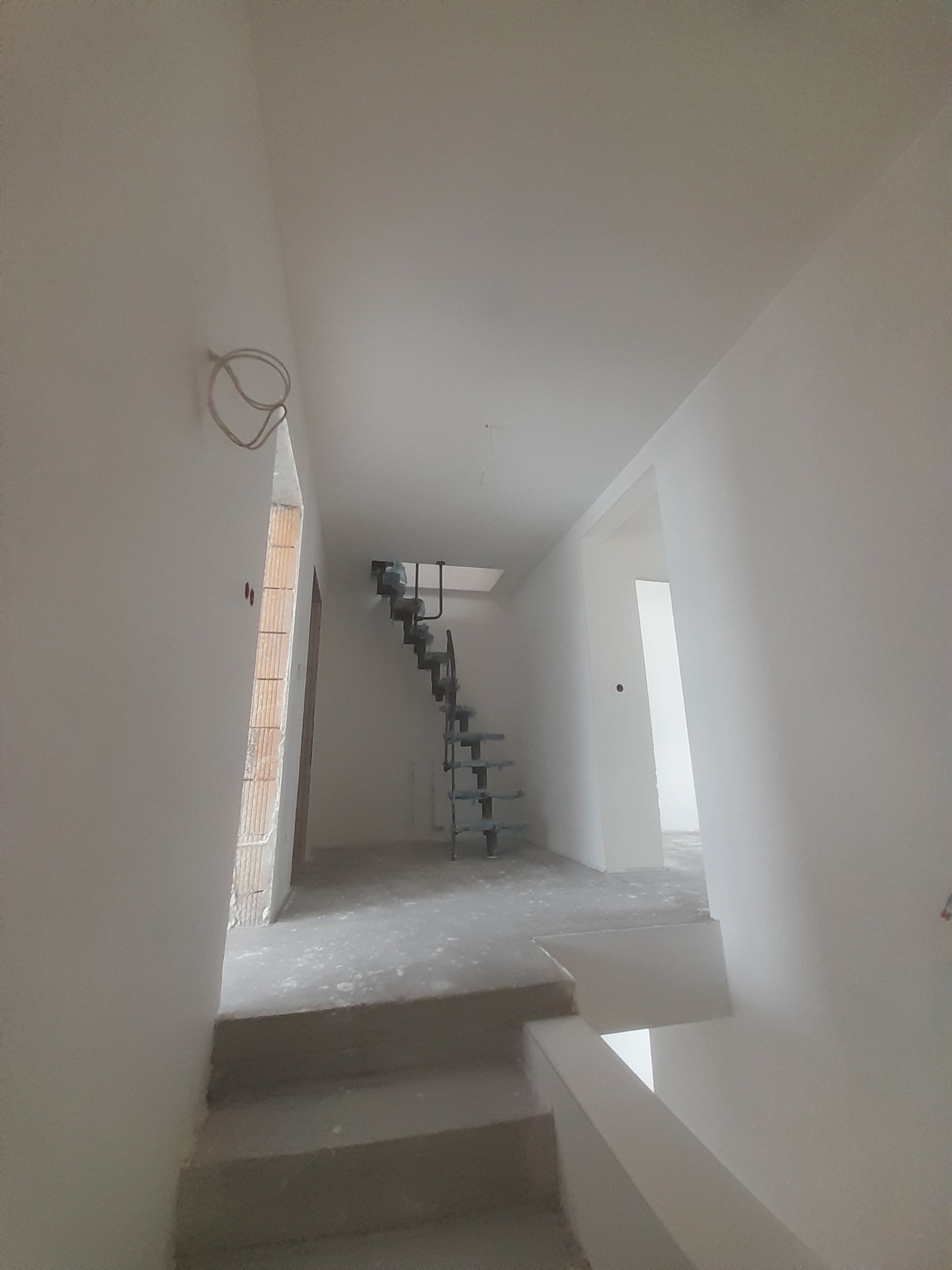 Wnętrze domu w trakcie prac wykończeniowych; widoczne surowe ściany, schody betonowe oraz metalowa konstrukcja schodów na piętro; przewody elektryczne wystające ze ściany.