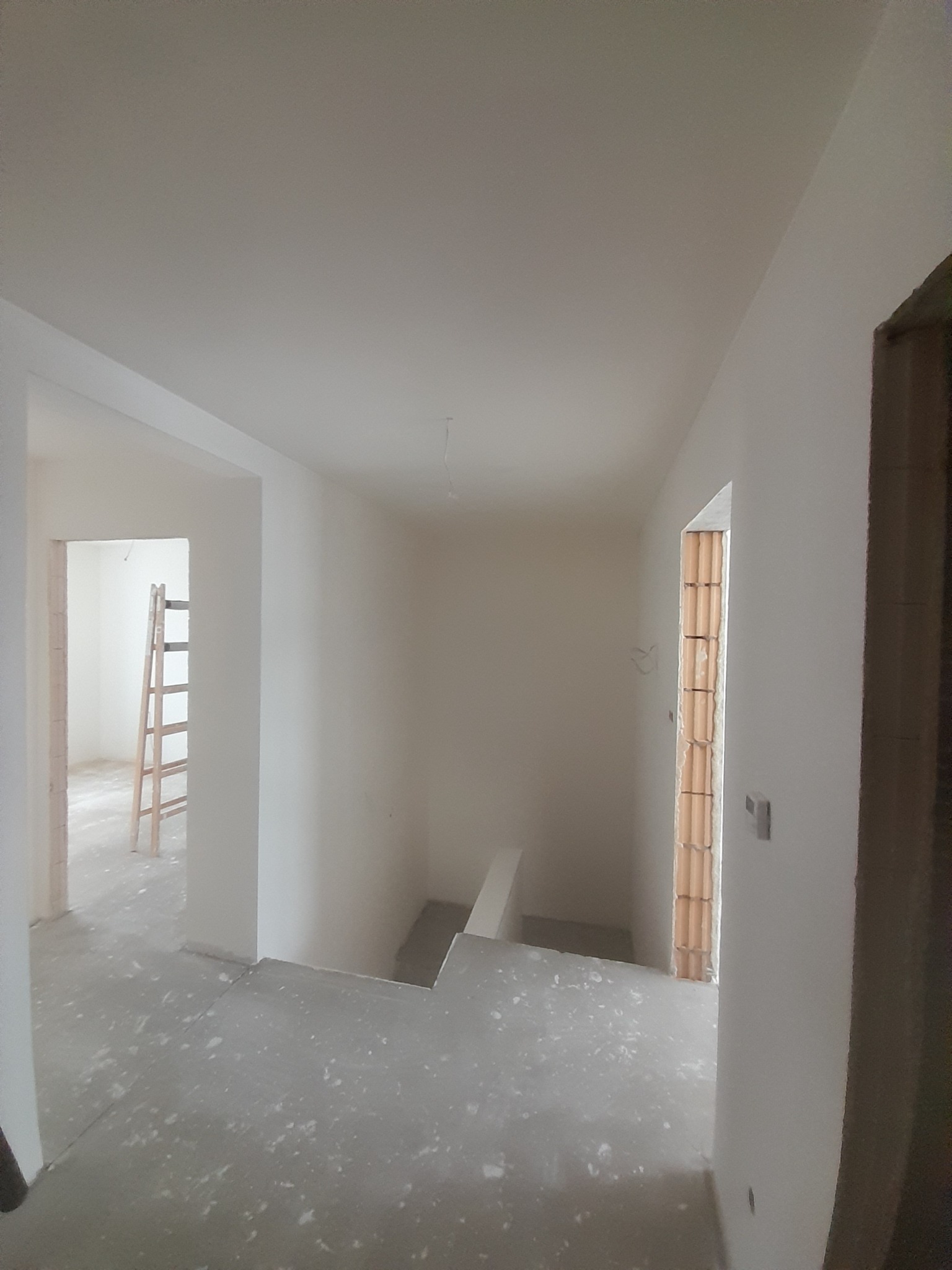 Surowe wnętrze w trakcie prac wykończeniowych: gładkie, białe ściany, szary betonowy podkład, widoczne cegły w ościeżnicy okiennej, drewniana drabina w tle.