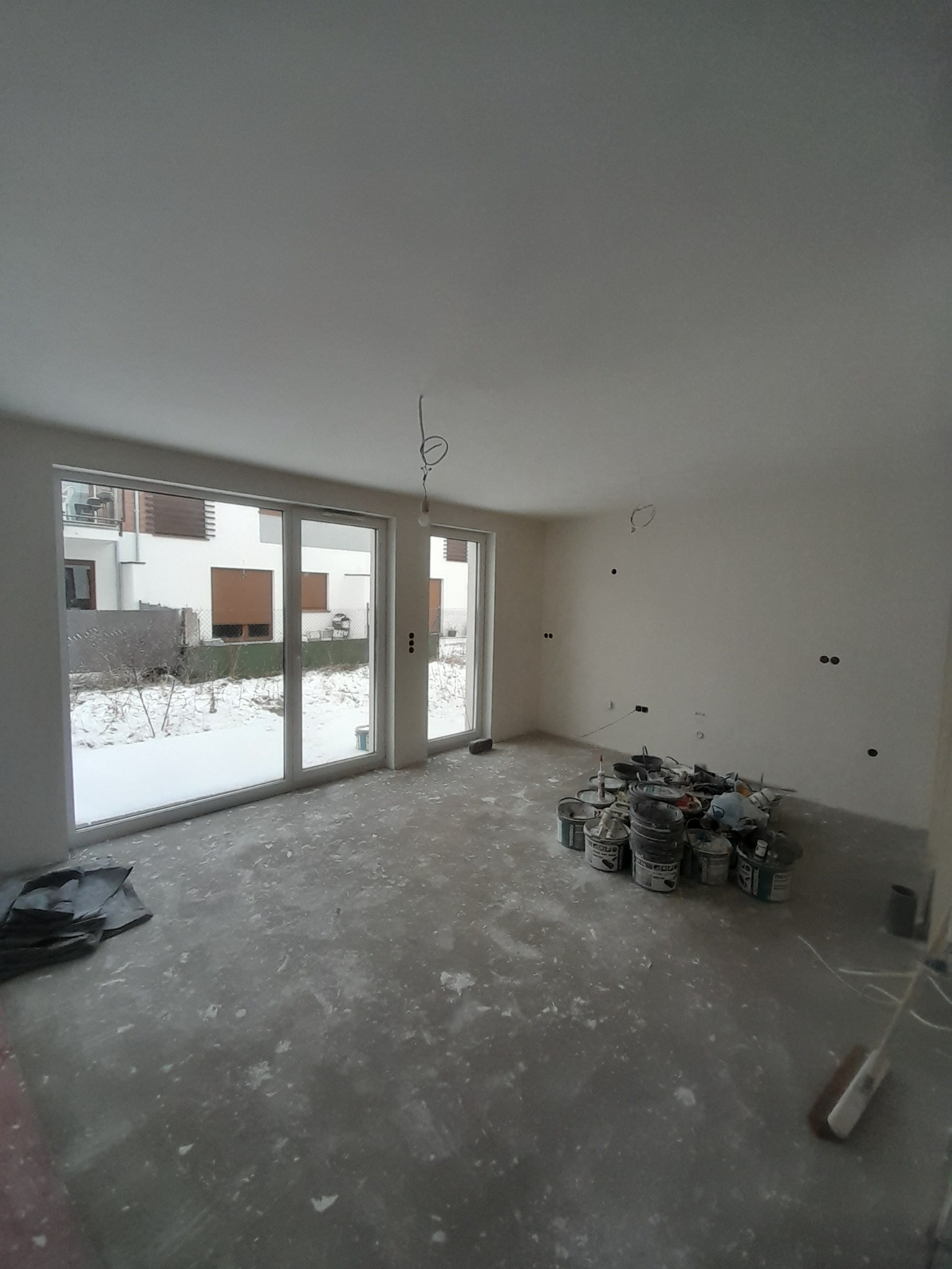 Wnętrze w trakcie prac wykończeniowych: widoczne puszki z farbą, zakurzona podłoga i ściany po wstępnym malowaniu, z widocznymi puszkami elektrycznymi. Na zewnątrz okien widoczny ośnieżony ogród.