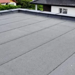 Szary, płaski dach z papy termozgrzewalnej na budynku jednorodzinnym, widoczne obróbki blacharskie i fragmenty innych budynków w tle.