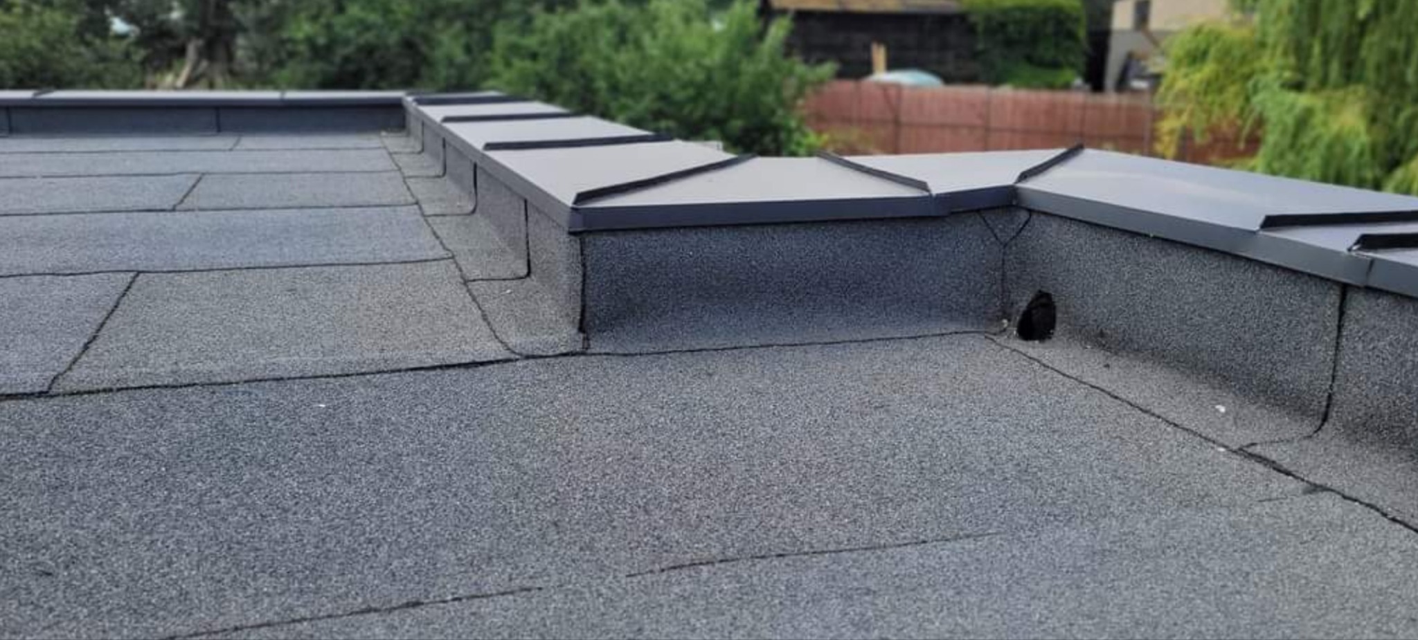 Szary dach pokryty papą z obróbkami blacharskimi w kolorze grafitowym, widoczne detale łączeń papy i otwór wentylacyjny, w tle zieleń drzew.
