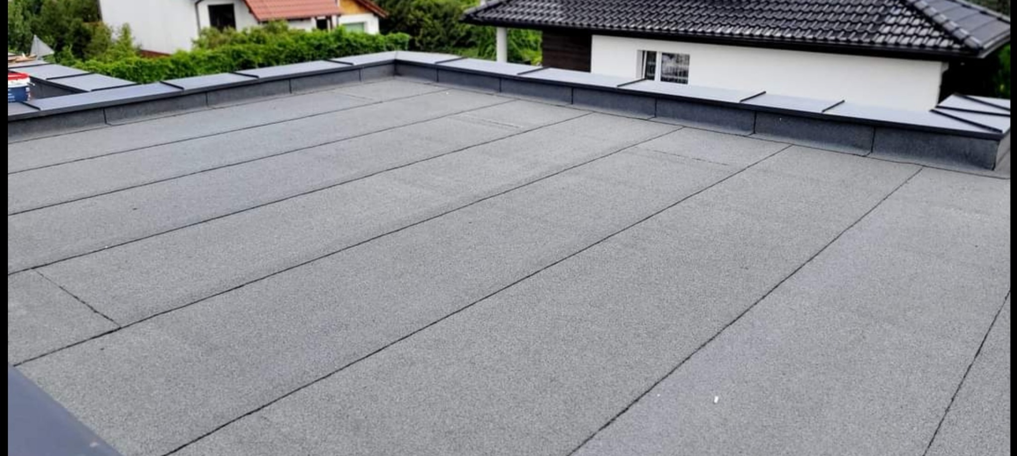 Szary, płaski dach z papy termozgrzewalnej na budynku jednorodzinnym, widoczne obróbki blacharskie i fragmenty innych budynków w tle.