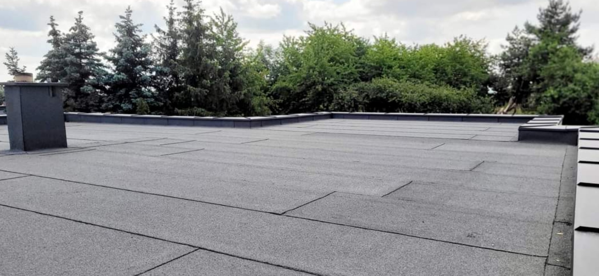 Płaski dach pokryty papą termozgrzewalną, widoczny komin i obróbki blacharskie, w tle drzewa iglaste i liściaste.