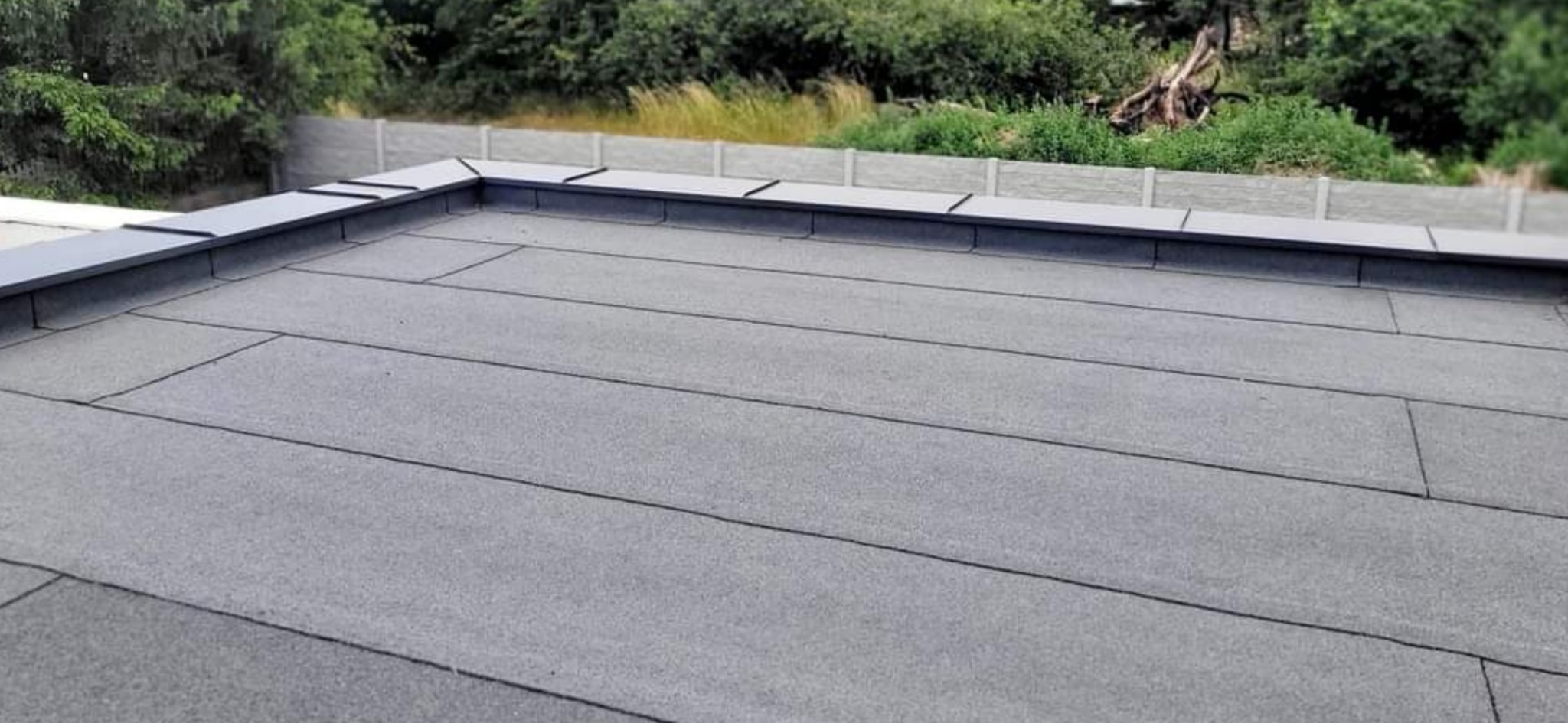 Płaski dach pokryty szarą papą termozgrzewalną z widocznymi łączeniami, zakończony szarym obrzeżem z blachy, na tle zieleni i betonowego ogrodzenia.
