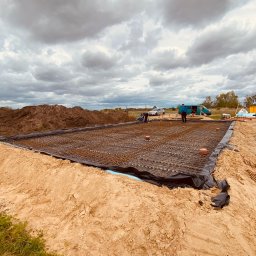 M&M Płyty Sp. Z o.o. - Zbrojenie płyty fundamentowej na czarnej folii, widoczne wystające pręty, pomarańczowe elementy dystansowe, dwóch pracowników w tle, hałda ziemi i pojazdy na działce budowlanej pod pochmurnym niebem.