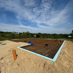 Przygotowany zbrojony szkielet fundamentów na piasku, obłożony niebieskim styropianem, z widocznymi rurami instalacyjnymi i shadow selfie na tle błękitnego nieba