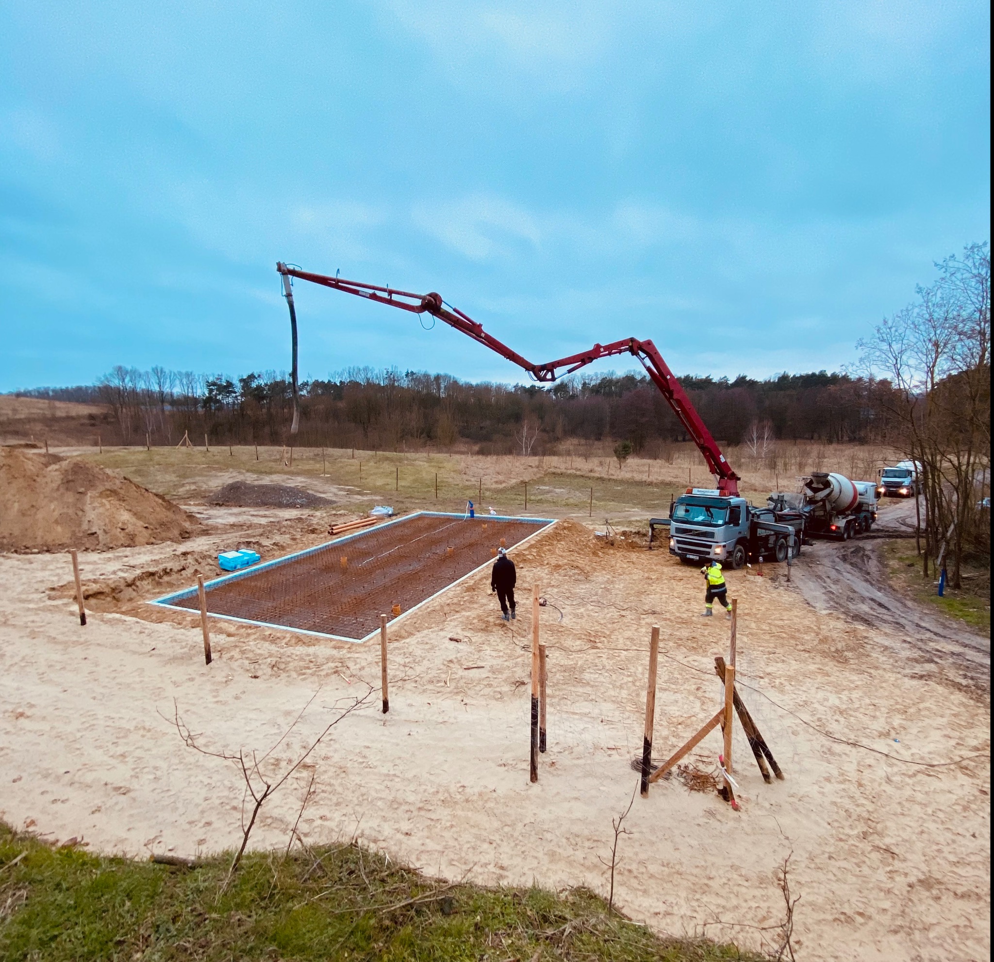 Wylewanie betonu do przygotowanego zbrojenia pod płytę fundamentową za pomocą pompy do betonu na tle piaszczystego terenu budowy i lasu.