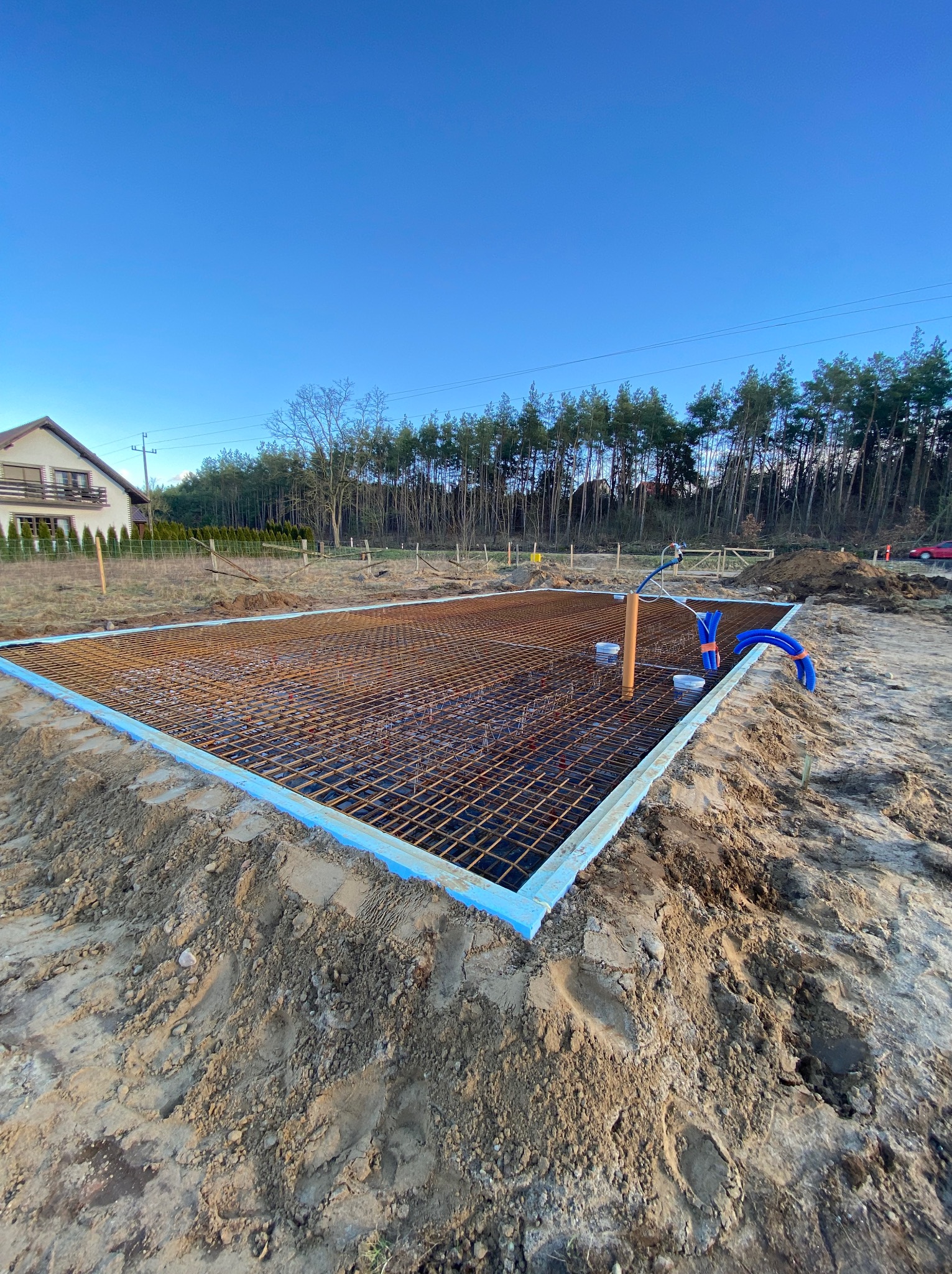 Przygotowany zbrojony szkielet fundamentu z izolacją obwodową, widoczne rury instalacyjne wystające z ziemi, w tle dom jednorodzinny i las.