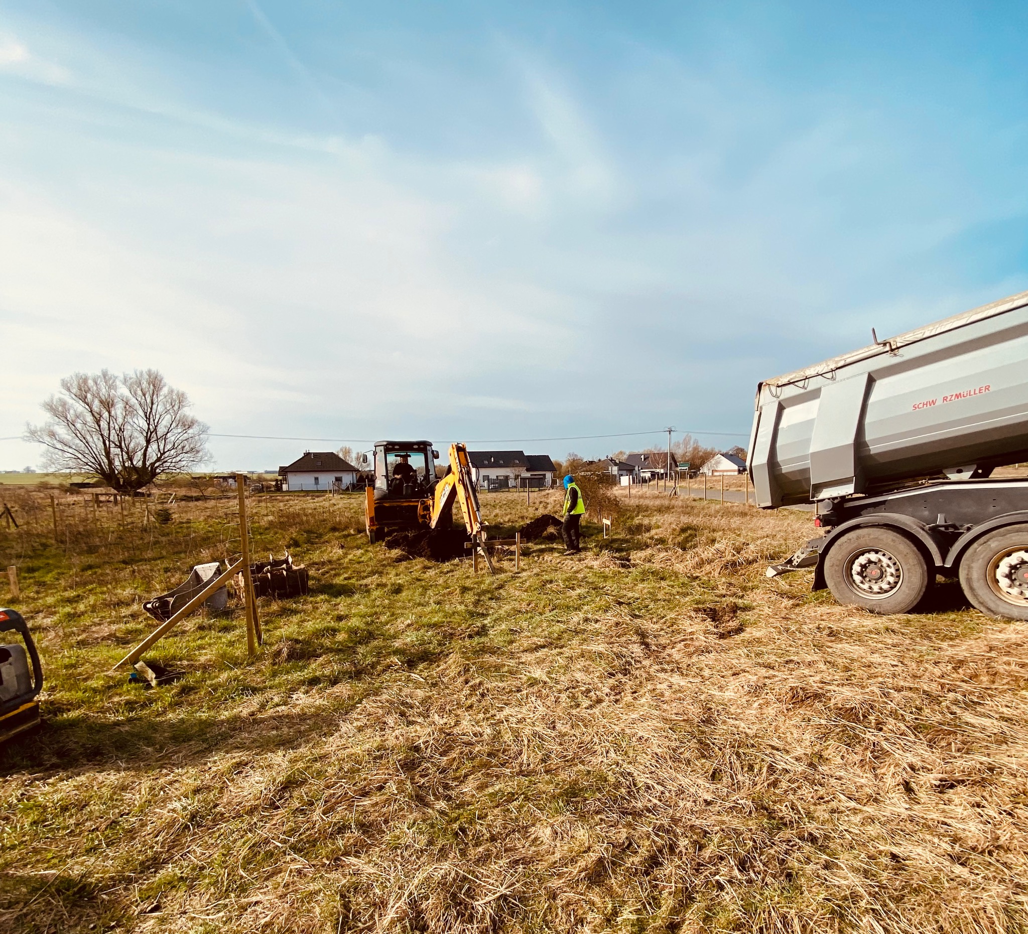 Mini koparka wykonuje wykop pod fundamenty, obok stoi wywrotka, a pracownik w kamizelce nadzoruje prace na tle zabudowań.