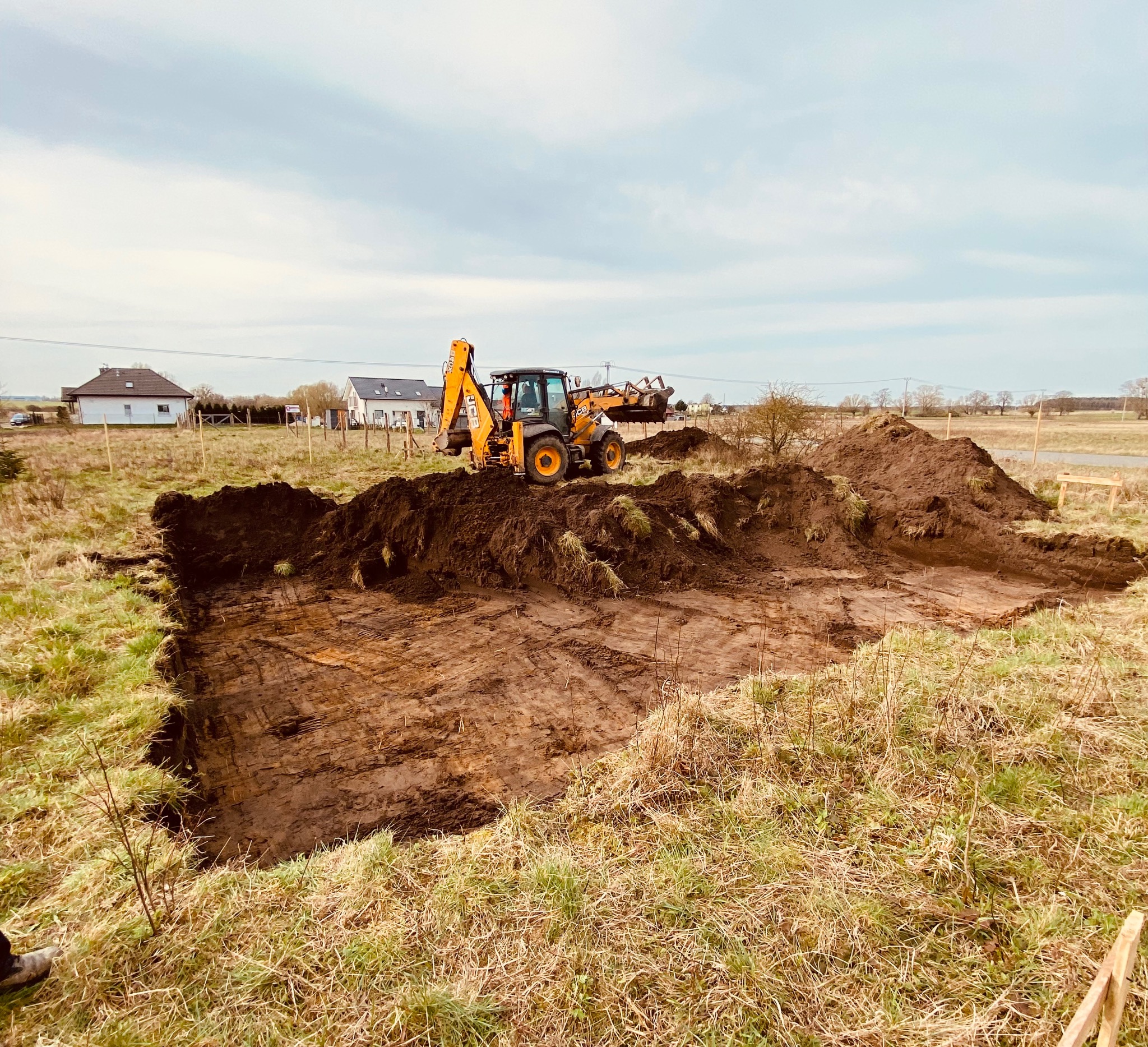 Żółta koparko-ładowarka JCB na placu budowy podczas prac ziemnych, wykop pod fundamenty w kształcie prostokąta, na obrzeżach trawa i drewniane słupki wyznaczające teren, w tle dwa domy jednorodzinne.