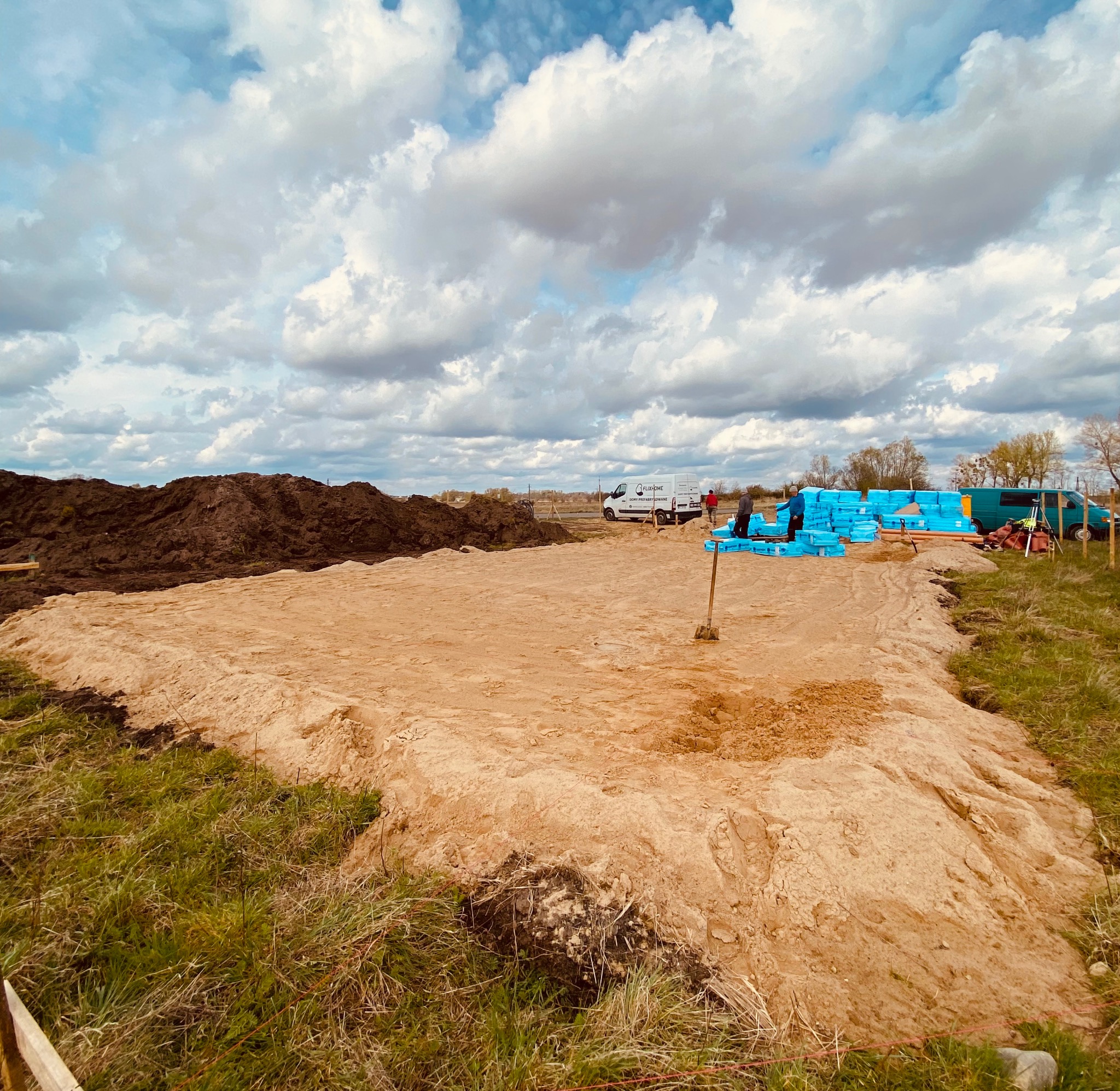Przygotowany teren pod budowę z wykopanym piaskiem, widoczne materiały izolacyjne i firmowy bus w tle.
