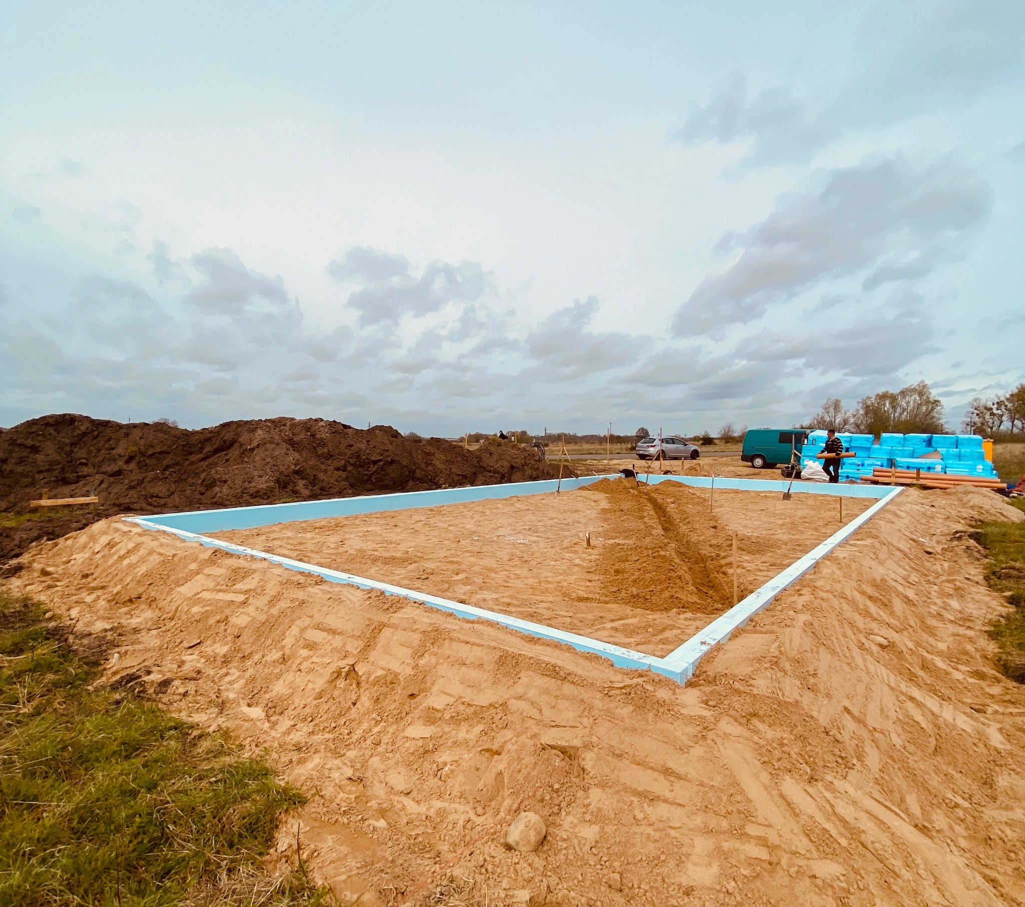 Przygotowany wykop pod fundamenty, obrysowany niebieskim styropianem, z widocznym stosem izolacji i pracownikiem w tle, na tle pochmurnego nieba.